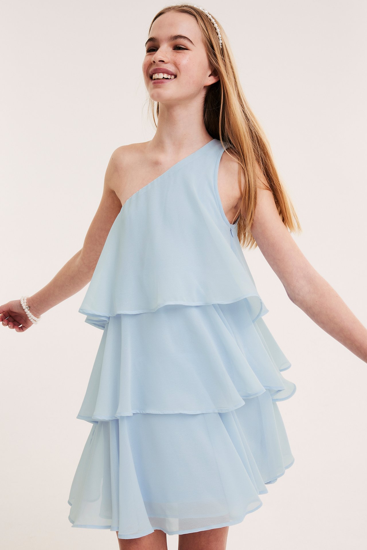 Framifrån, ett barn i ljusblå enaxlad klänning med volanger, ett pärldiadem och ett pärlarmband.