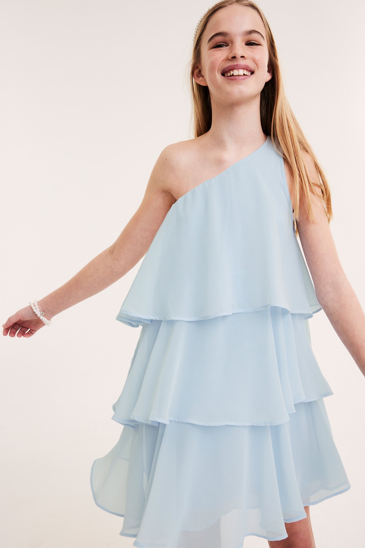 Frontvy: Barn i en ljusblå enaxlad klänning med volanger, glittrande diadem och pärlarmband.