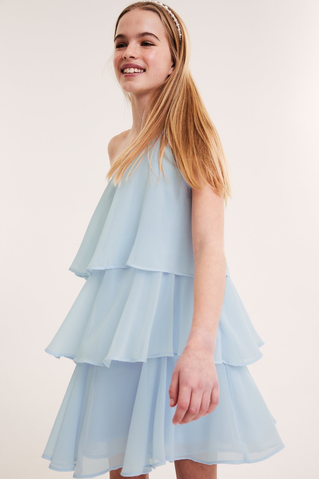 Sidovy på ett leende barn i en ljusblå one-shoulder klänning med volanger och pärlband.