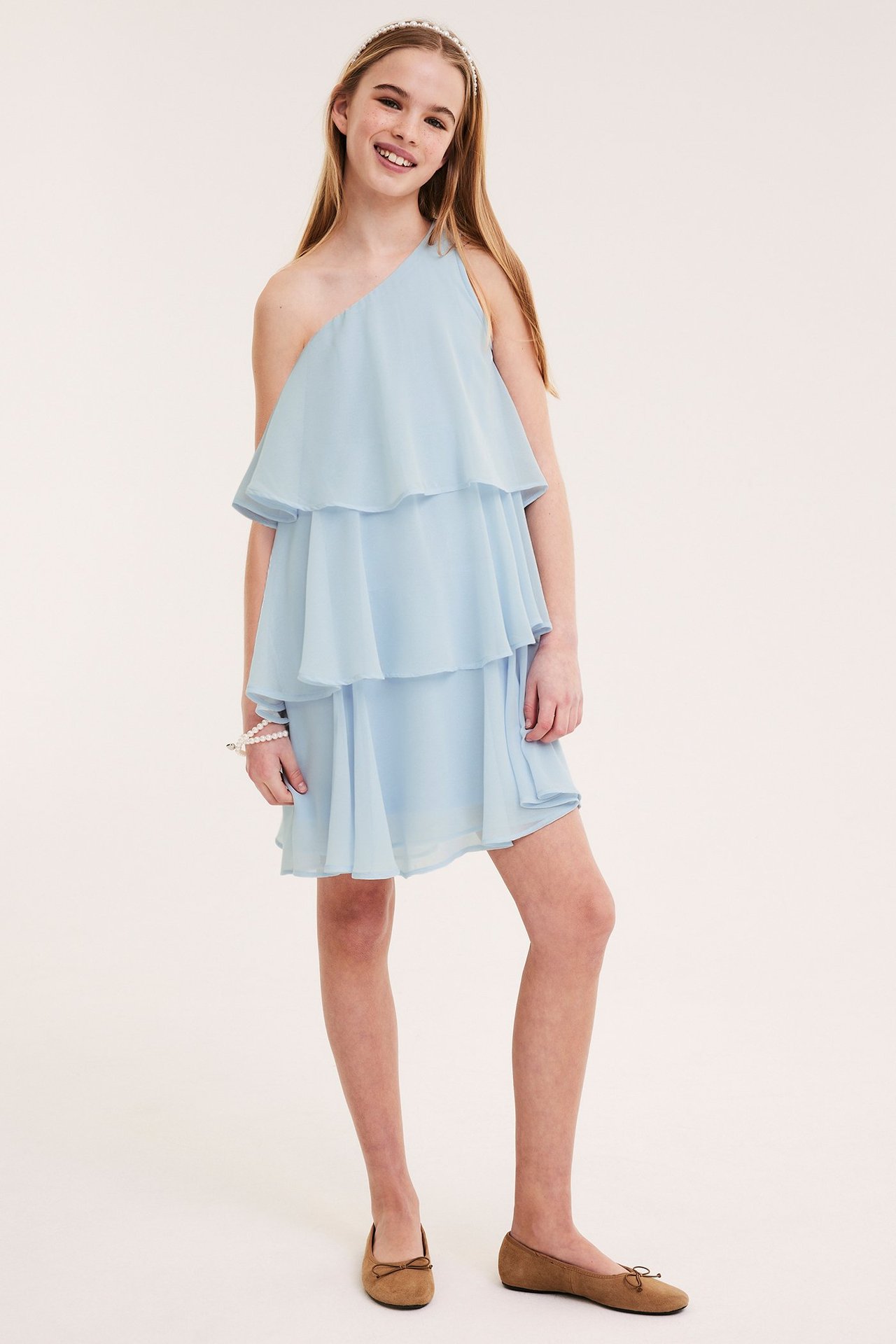 Frontvy av ett barn i ljusblå one-shoulder volangklänning, pärlband, pärlarmband och bruna ballerinaskor.