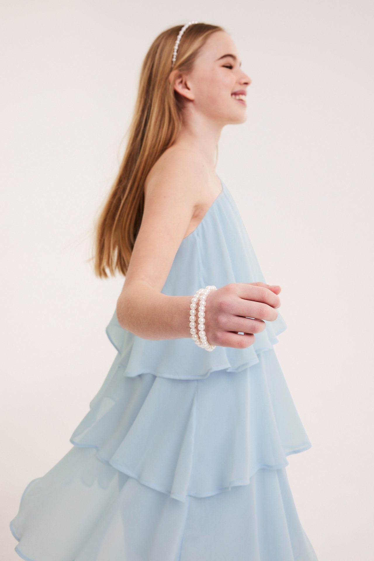 Widok z boku dziecka w jasnoniebieskiej sukience na jedno ramię z falbanami, opasce z pereł i dwóch bransoletkach.