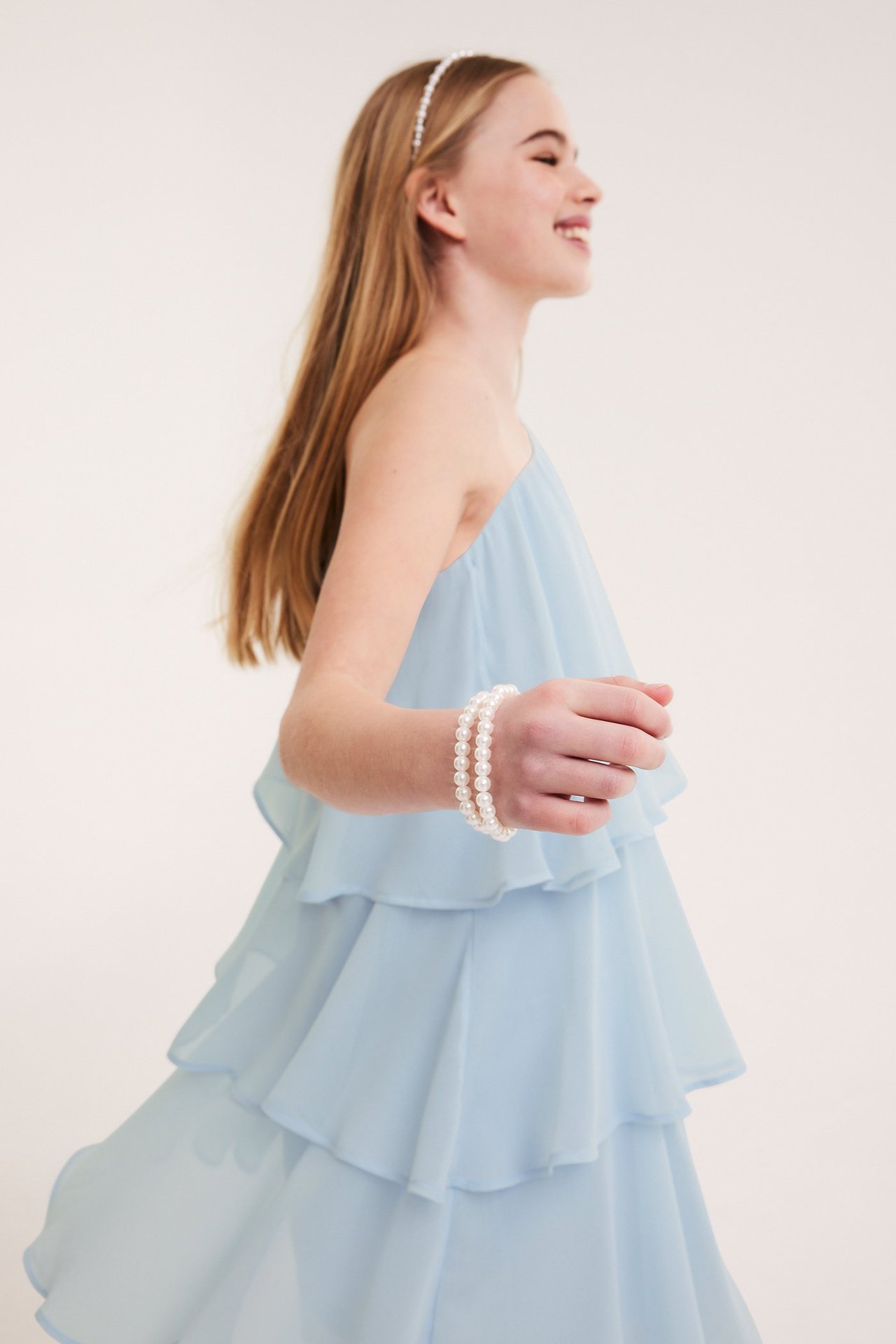 Widok z boku dziecka w jasnoniebieskiej sukience na jedno ramię z falbanami, opasce z pereł i dwóch bransoletkach.