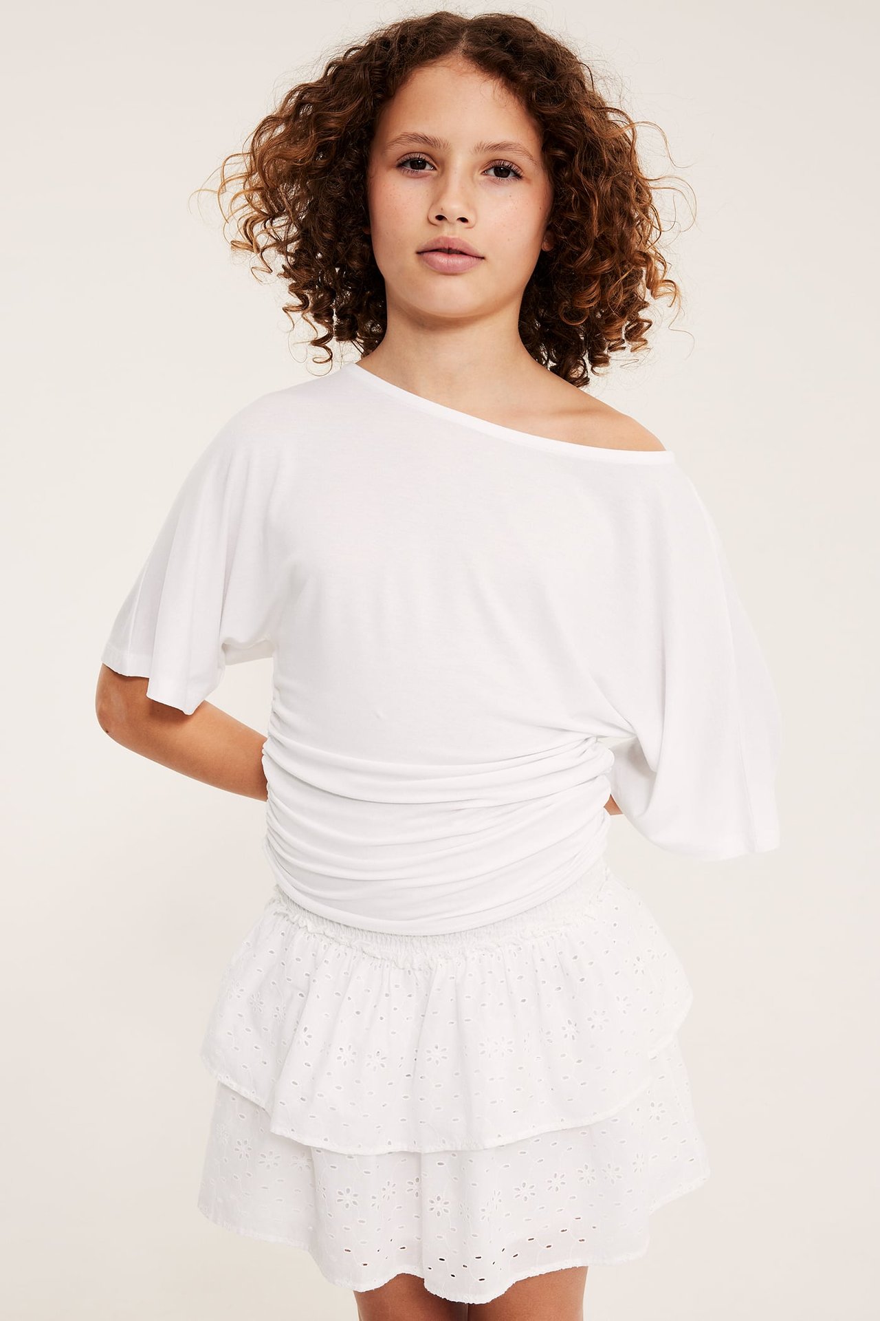 Forfra: Barn i hvitt kort broderie anglaise-skjørt med smockmidje og en hvit off-shoulder topp.