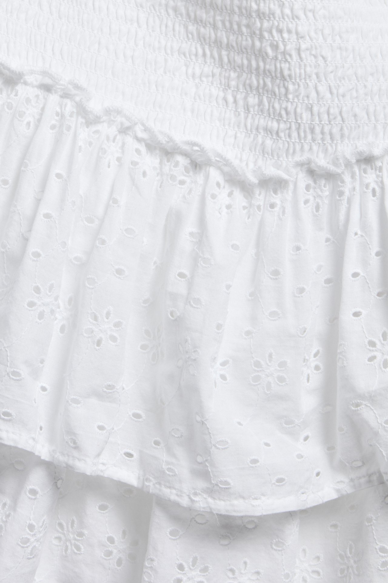 Närbild av en kort vit kjol i broderie anglaise med smockad midja och volanger.