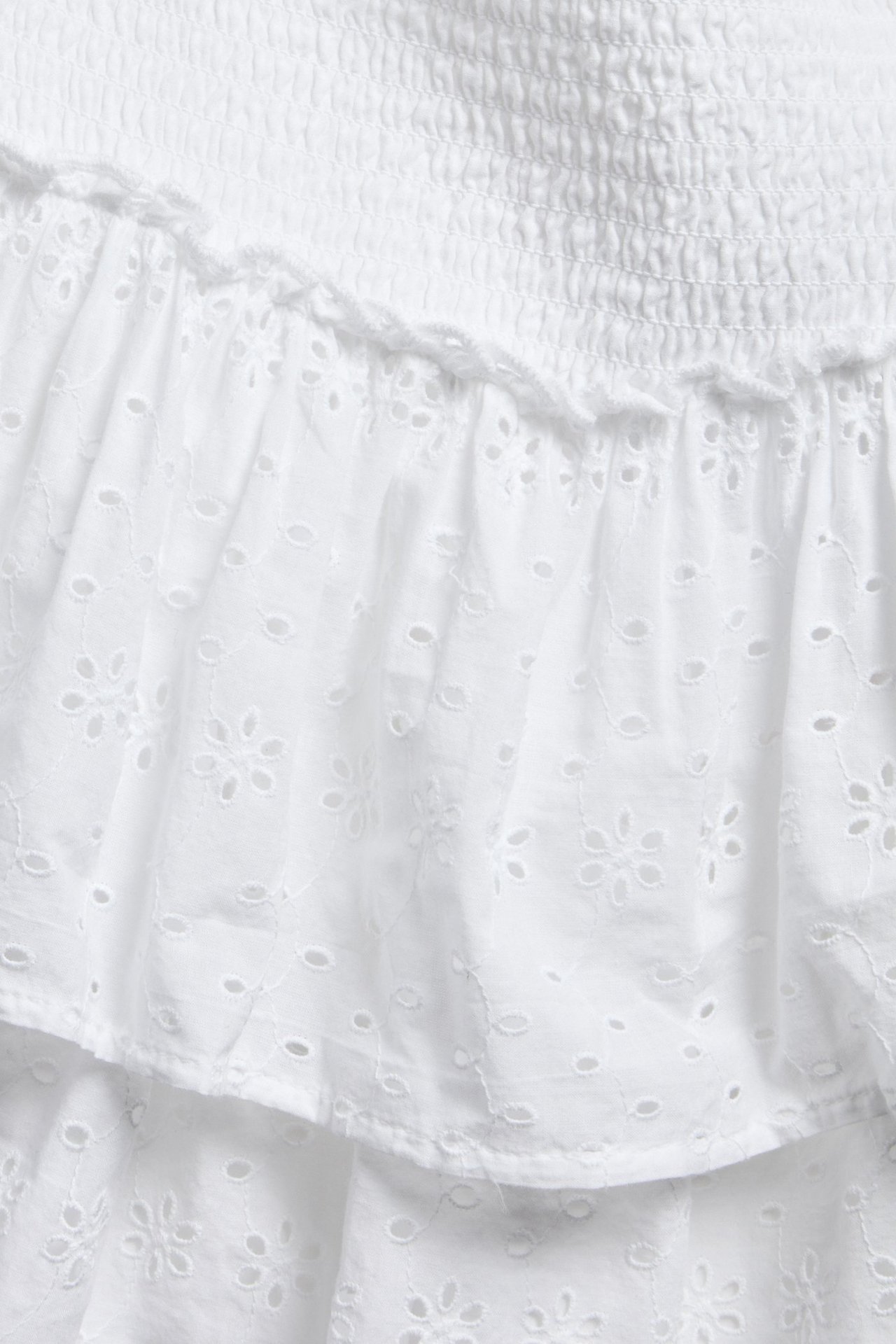 Närbild av en kort vit kjol i broderie anglaise med smockad midja och volanger.