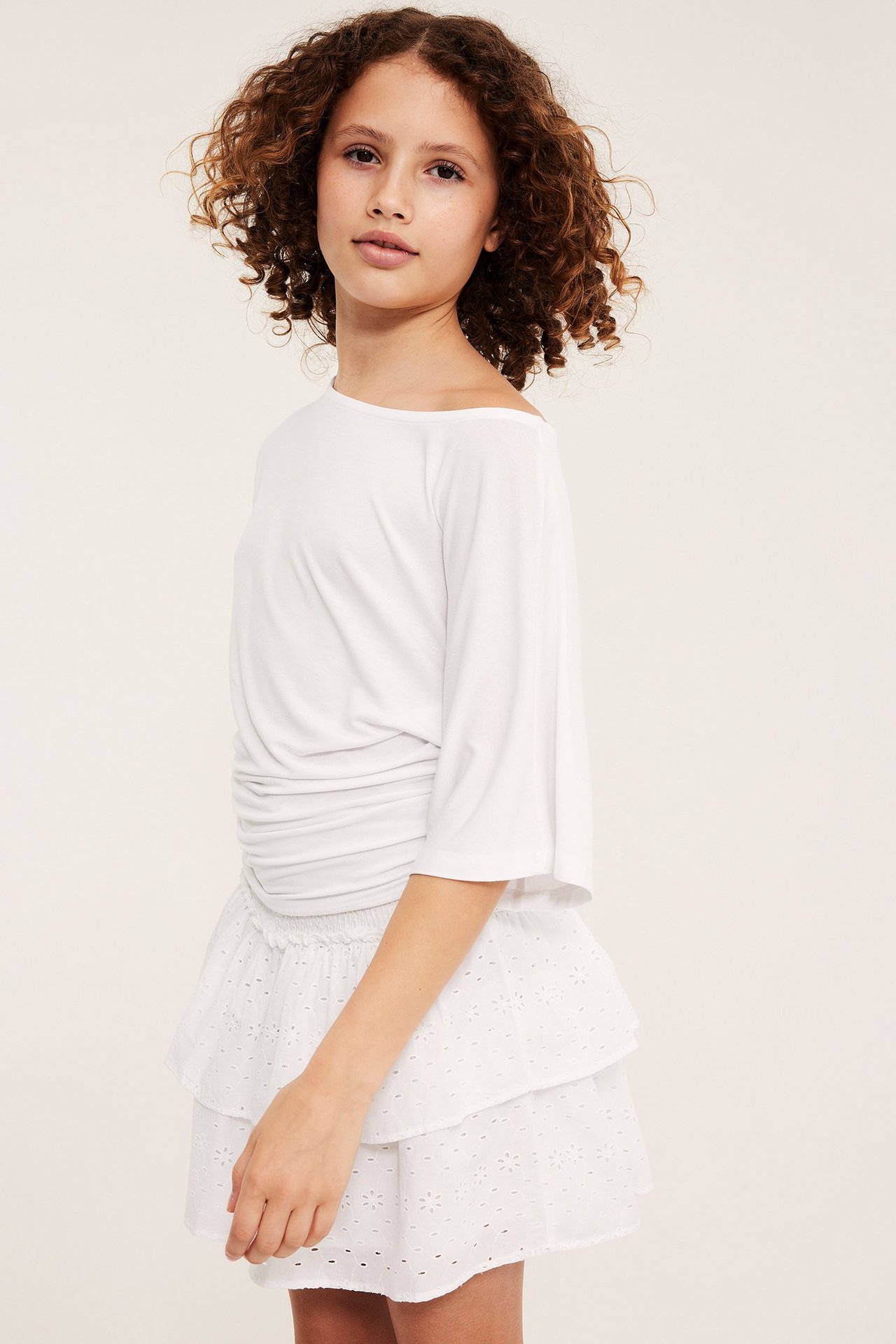 Framifrån: ett barn i vit broderie anglaise kort kjol med volanger och smock i midjan, och en vit topp.