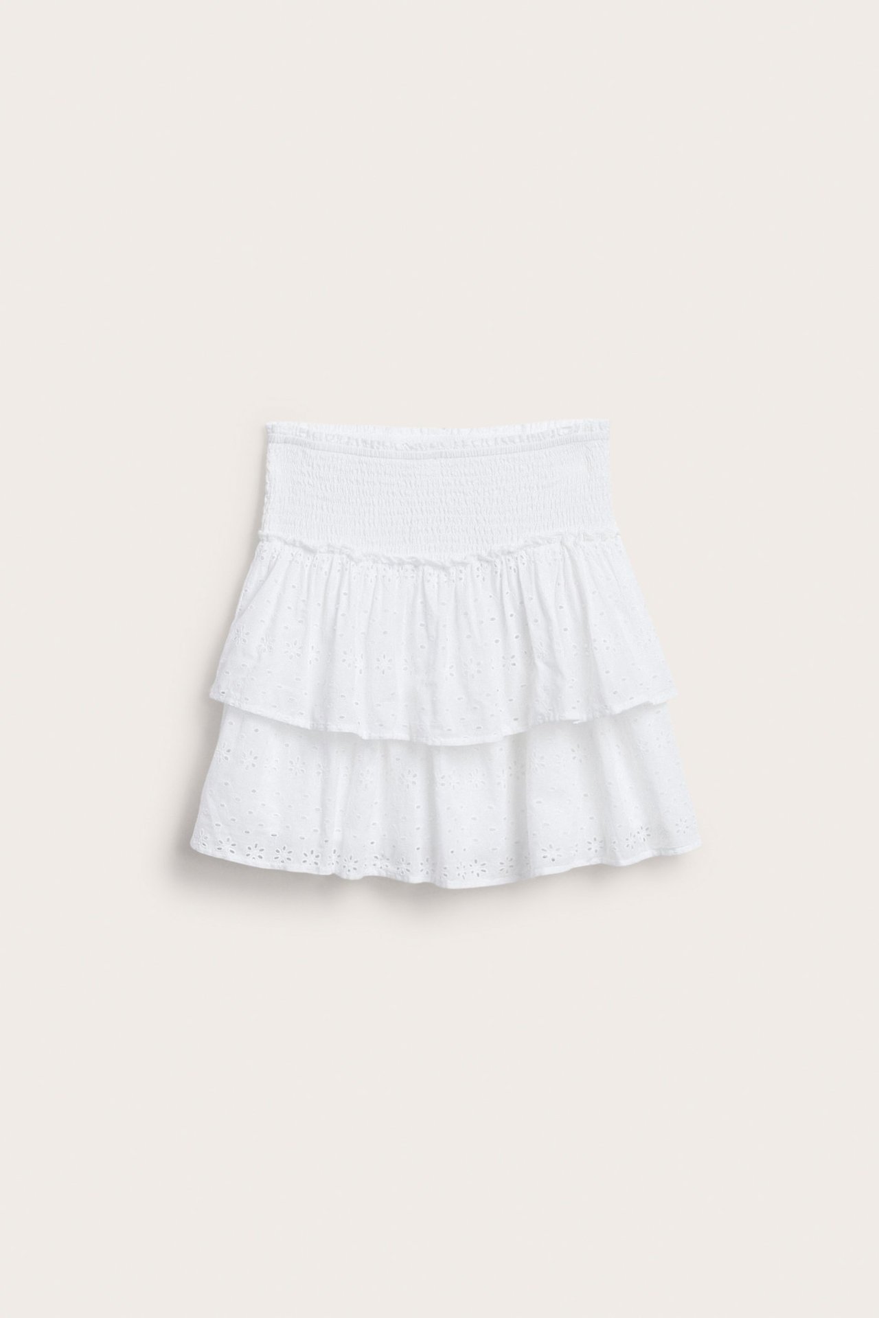 Framifrån, en vit kort barnkjol med smock i midjan och broderie anglaise-mönster i två volanger.