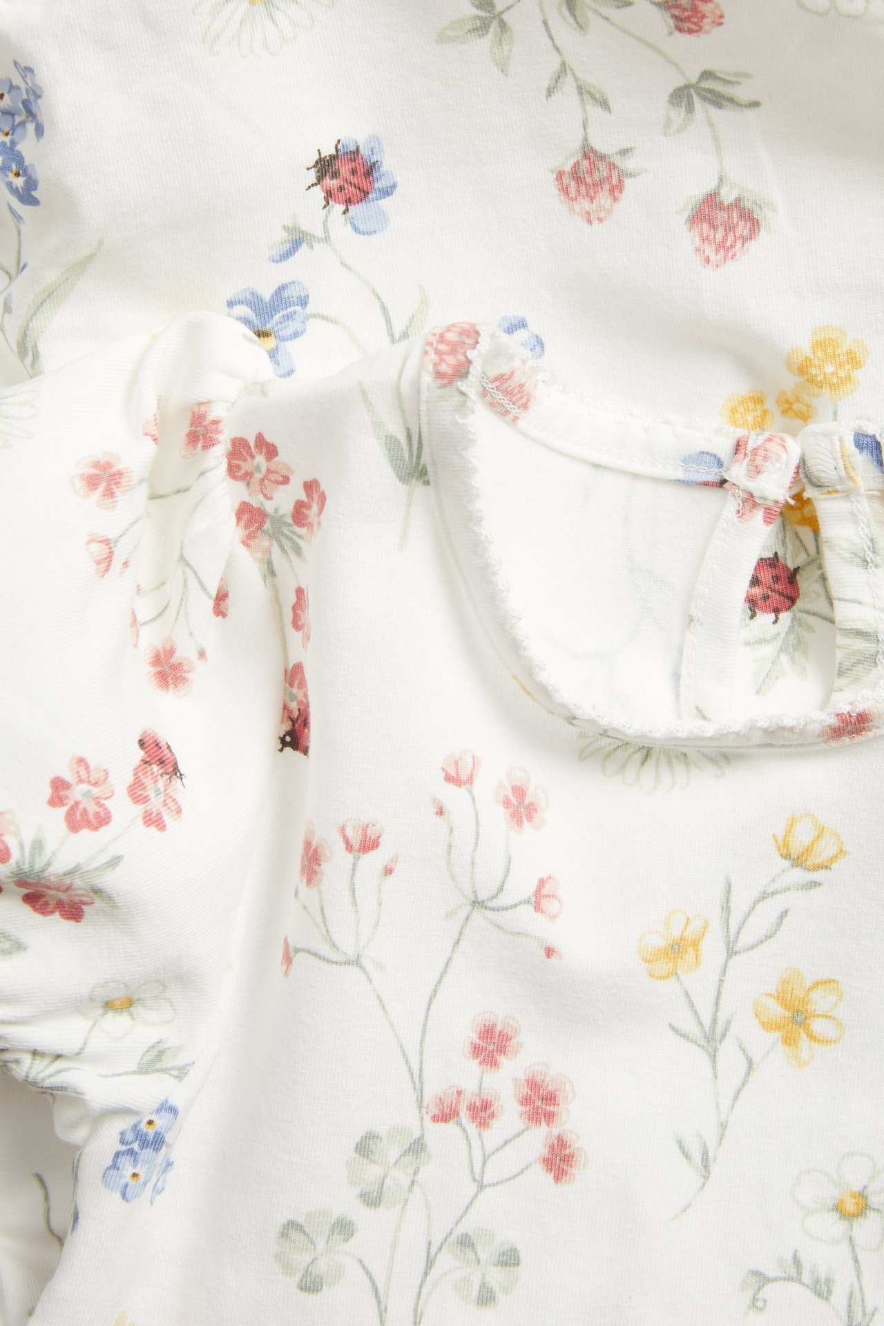 Lähikuva luonnonvalkoisesta vauvanbodysta, jossa kukka- ja leppäkerttakuvio, lyhyet pussihihat ja picot-reunus.