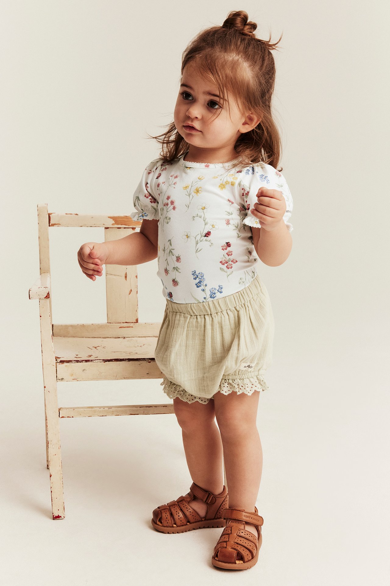 Frontvisning av baby i en offwhite blomstret kortermet body, lysebeige shorts med blonde, og brune sandaler.