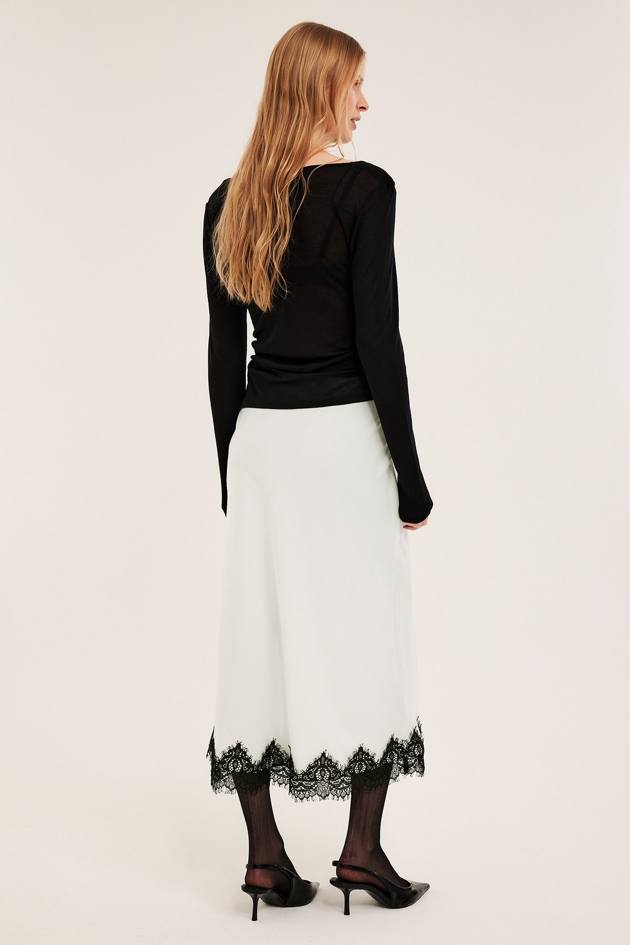 Bakfra: kvinne i offwhite satengskjørt med svart blondekant, svart topp, strømpebukser og høyhælte sko.