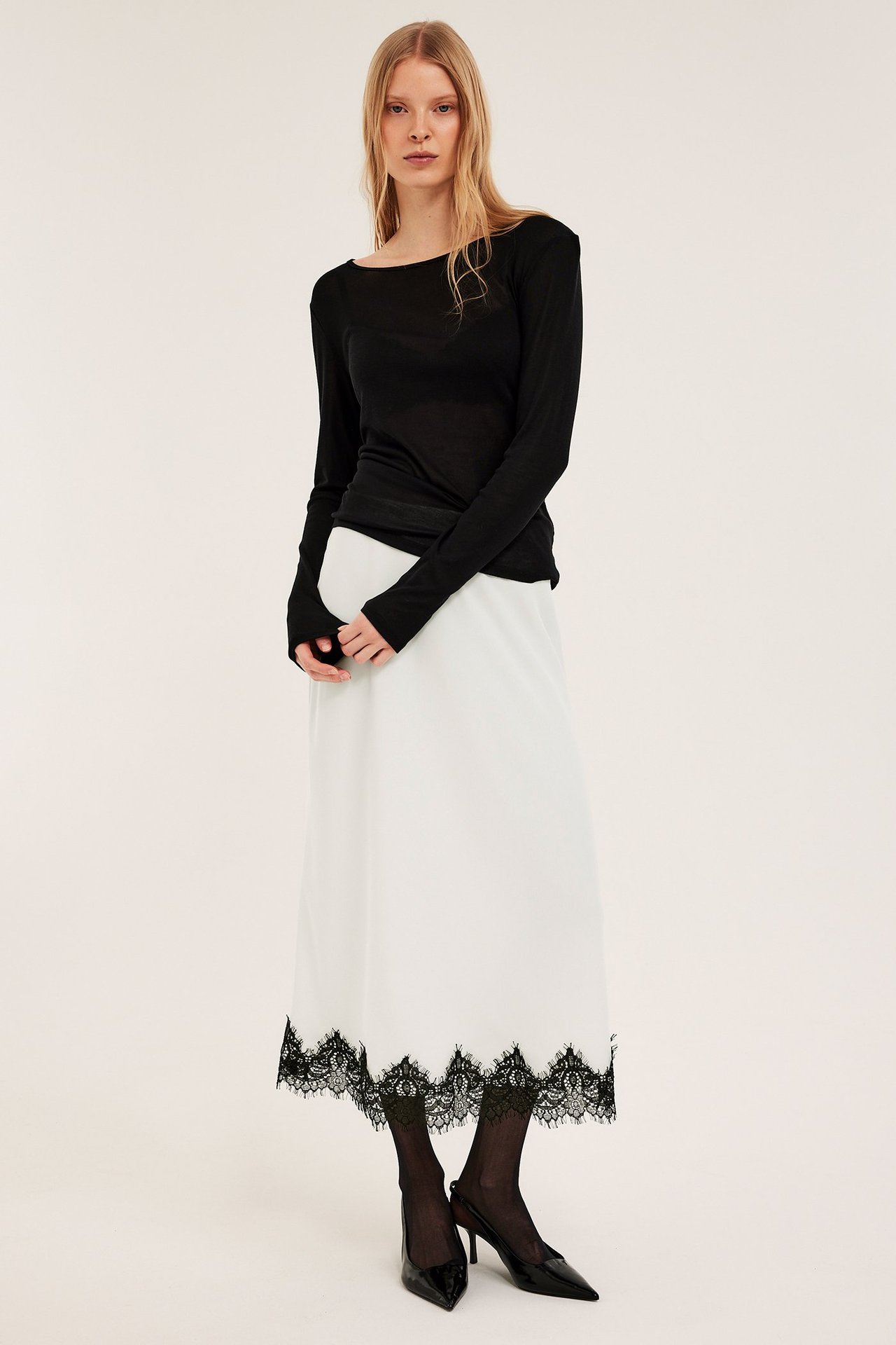 Kvinna i offwhite satinkjol med svart spetskant, svart långärmad topp, svarta strumpbyxor och klackskor.