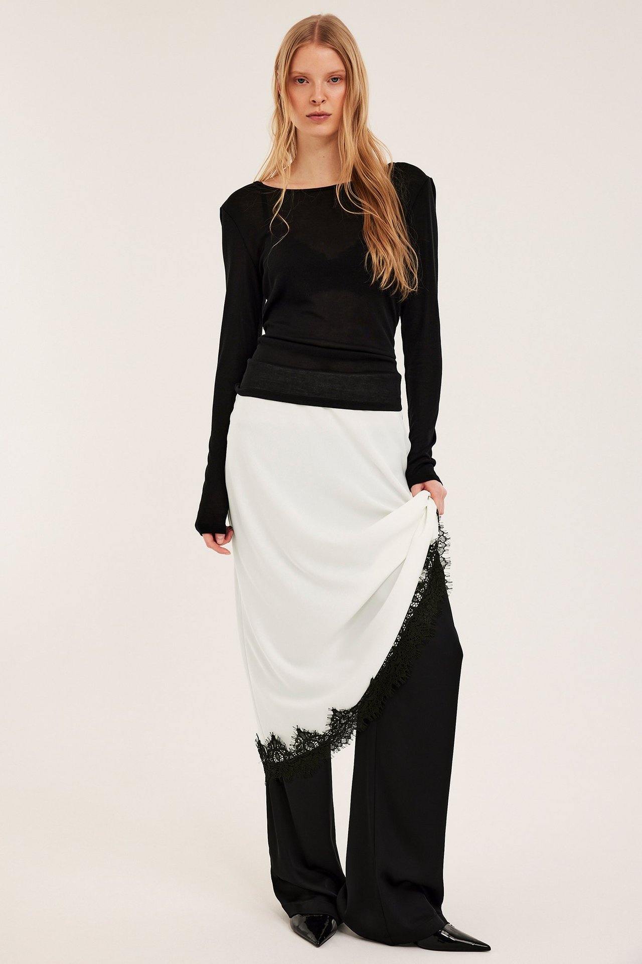Widok z przodu: kobieta trzyma ecru satynową spódnicę z czarną koronką na czarnym topie i spodniach.