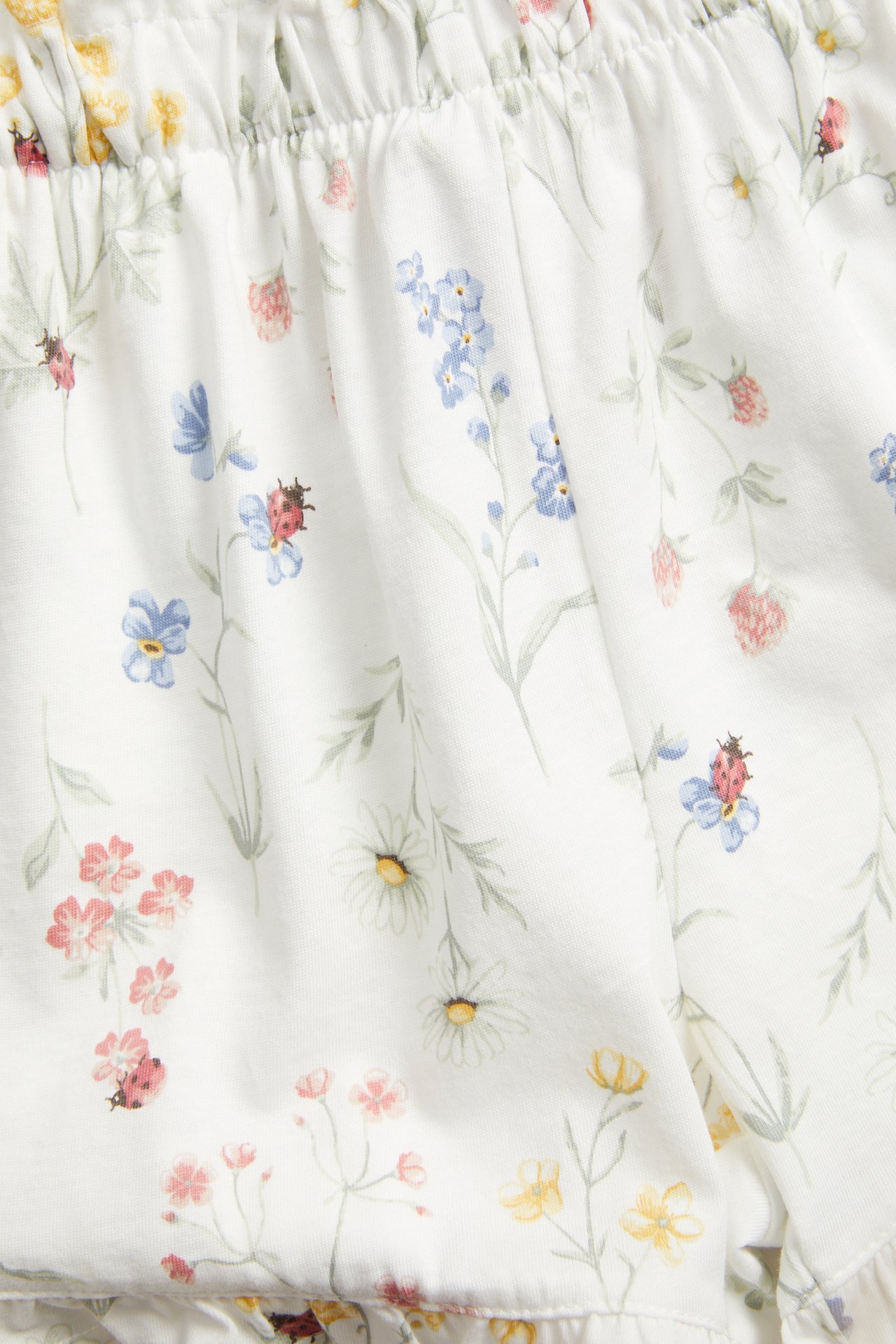 Närbild av offwhite bomullsjersey-shorts med blommigt och nyckelpigemönster, med resår i midjan.