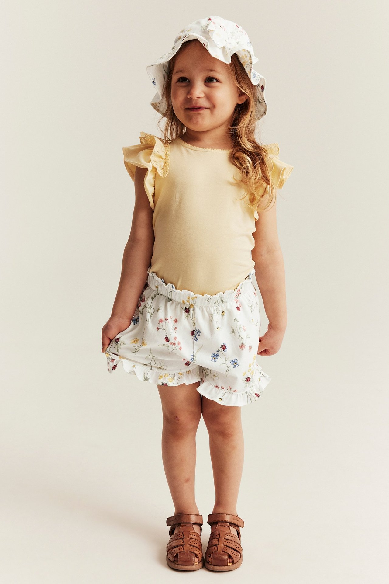 Frontal utsikt: Barnet har på seg offwhite blomstrete jersey shorts med volanger, gul topp, blomsterhatt og brune sandaler.