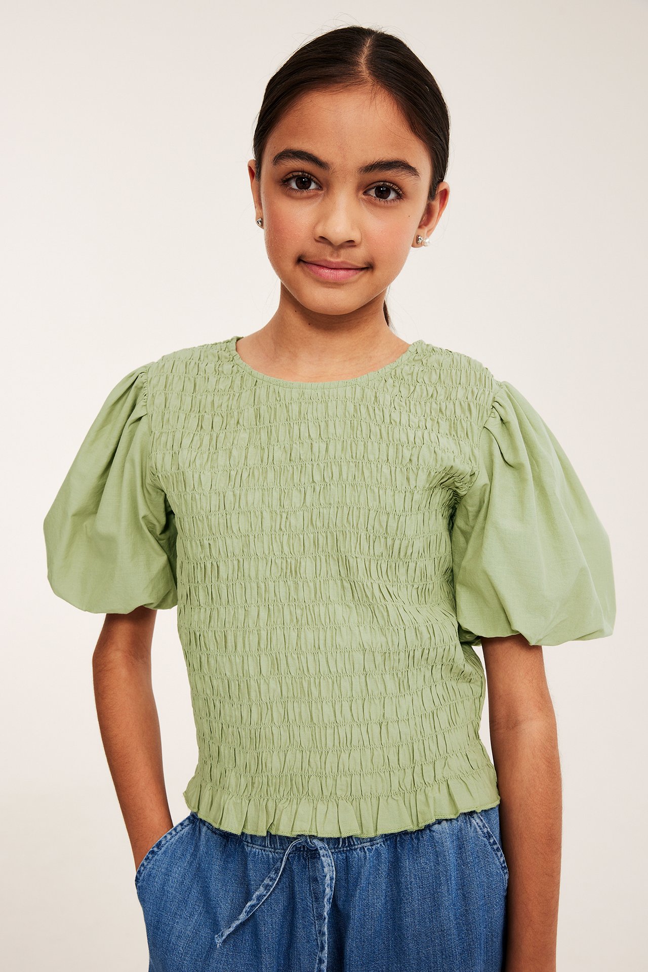 Dziecko nosi jasnozielony bawełniany top z marszczeniem, bufiastymi rękawami i niebieskie dżinsy.