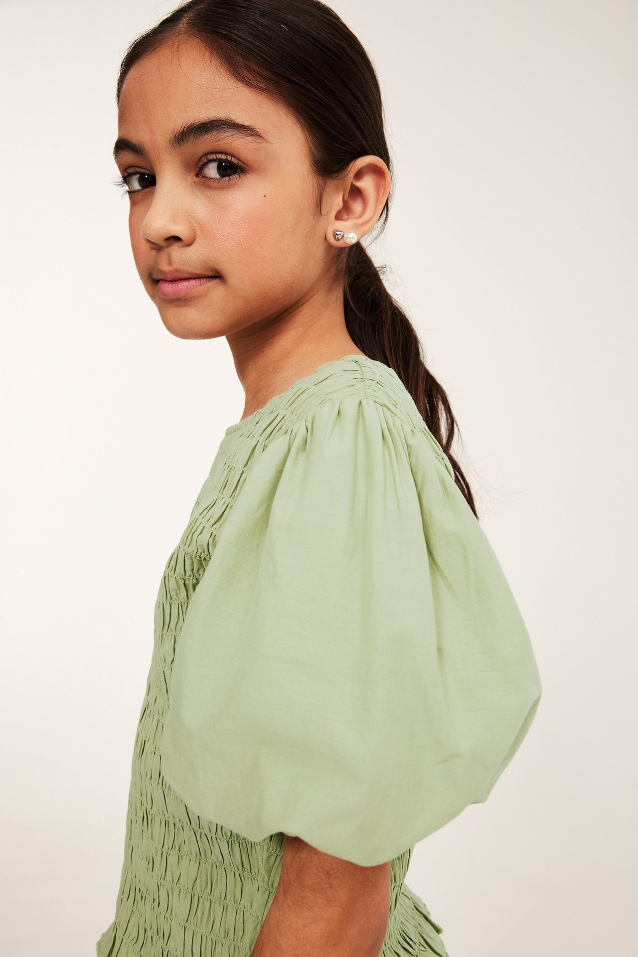 Zbliżenie dziewczynki w jasnozielonym topie ze smoczkowaniem, bufiastymi rękawami i kolczykami z pereł.