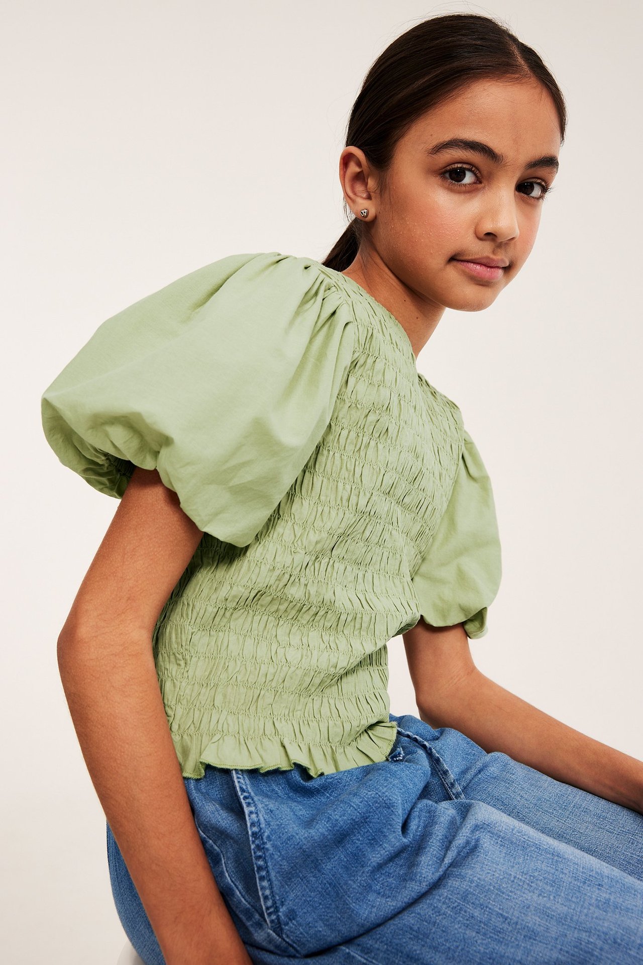 Edestäpäin lapsi yllään vaaleanvihreä puuvillatoppi, jossa smokkirypytys ja pöyhkeät hihat, sinisten farkkujen kanssa.