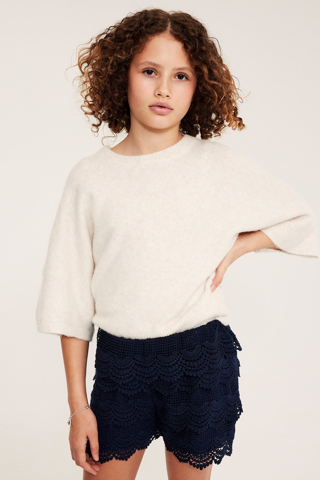 Edestä: Lapsi pukeutunut laivastonsinisiin pitsishortseihin ja löysään vaaleanbeesiin neuleeseen.