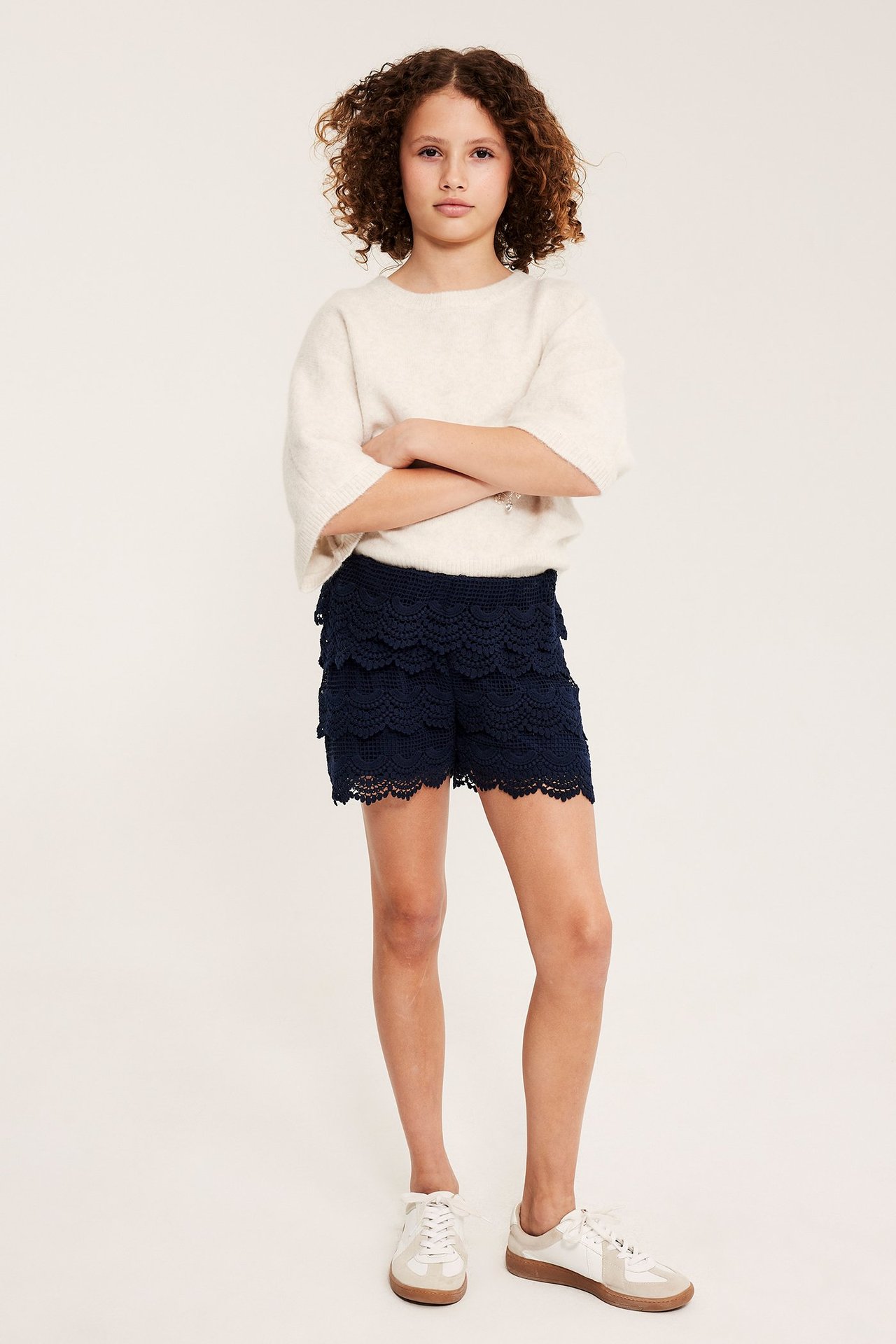 Widok z przodu, dziecko w granatowych koronkowych szortach z falbanami, jasnobeżowym swetrze i białych trampkach.