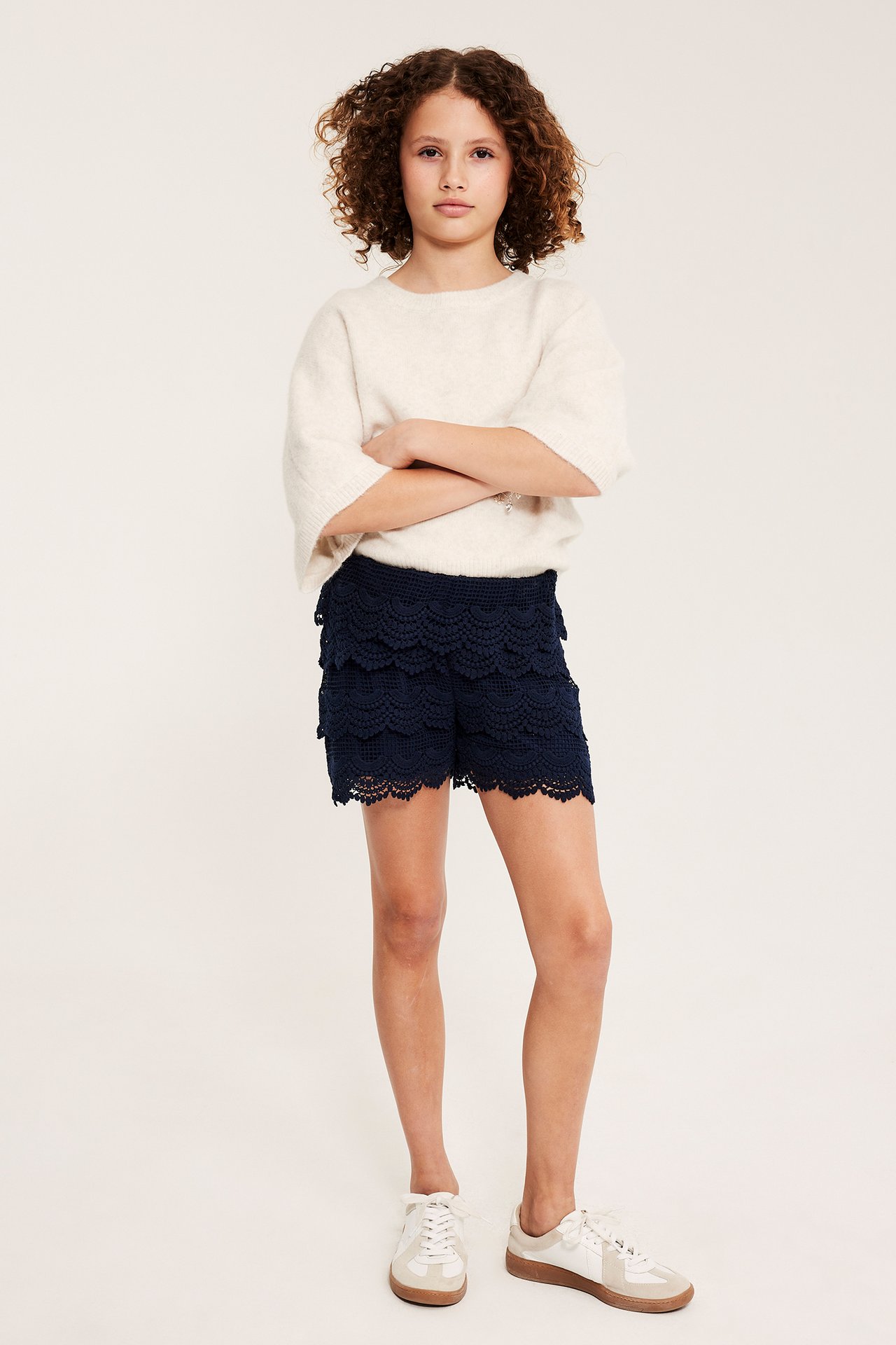 Frontutsikt, barn i marineblå volang-blonde shorts, en lys beige genser og hvite joggesko.