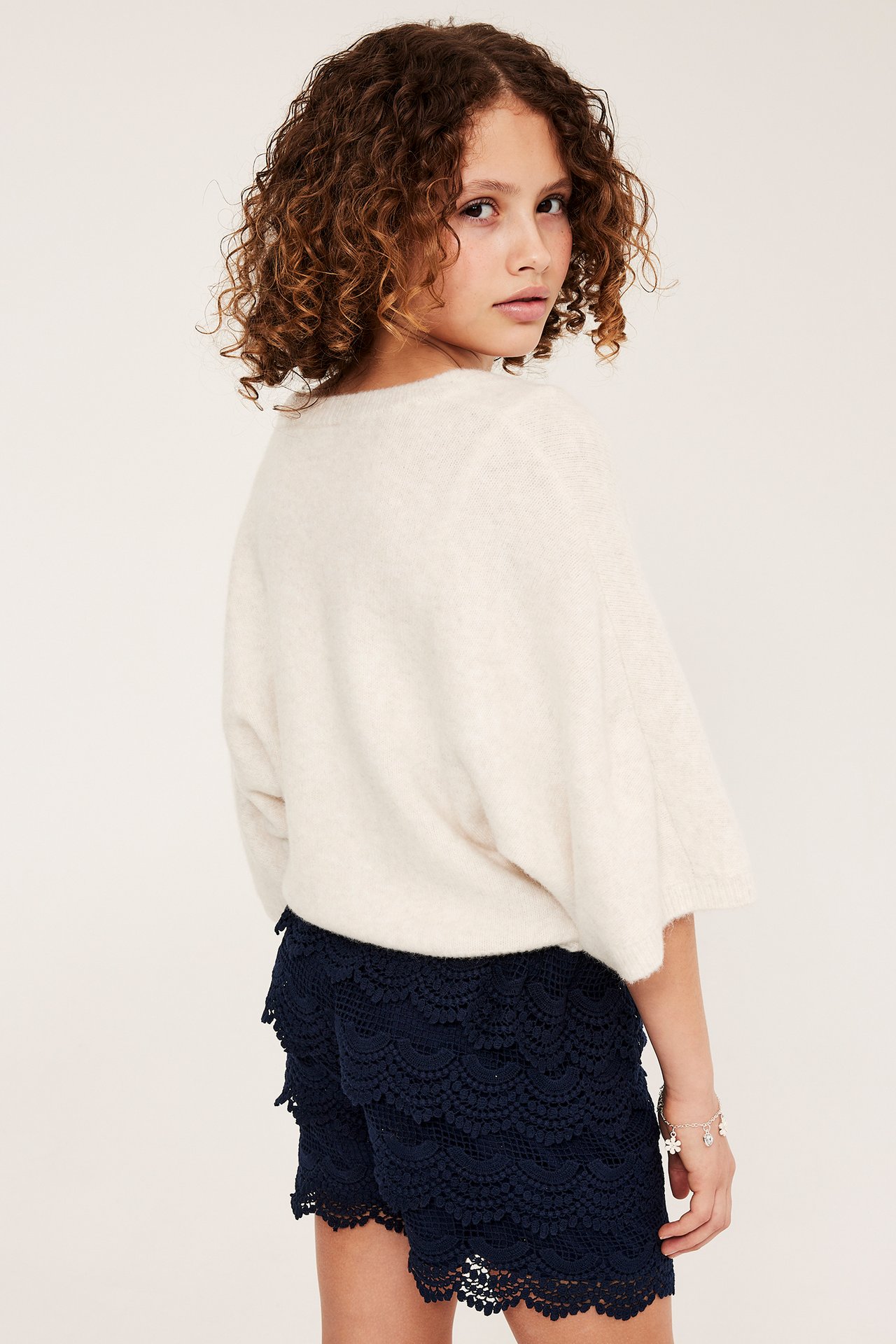 Widok z tyłu dziecka w granatowych koronkowych szortach, kremowym luźnym swetrze i bransoletce.