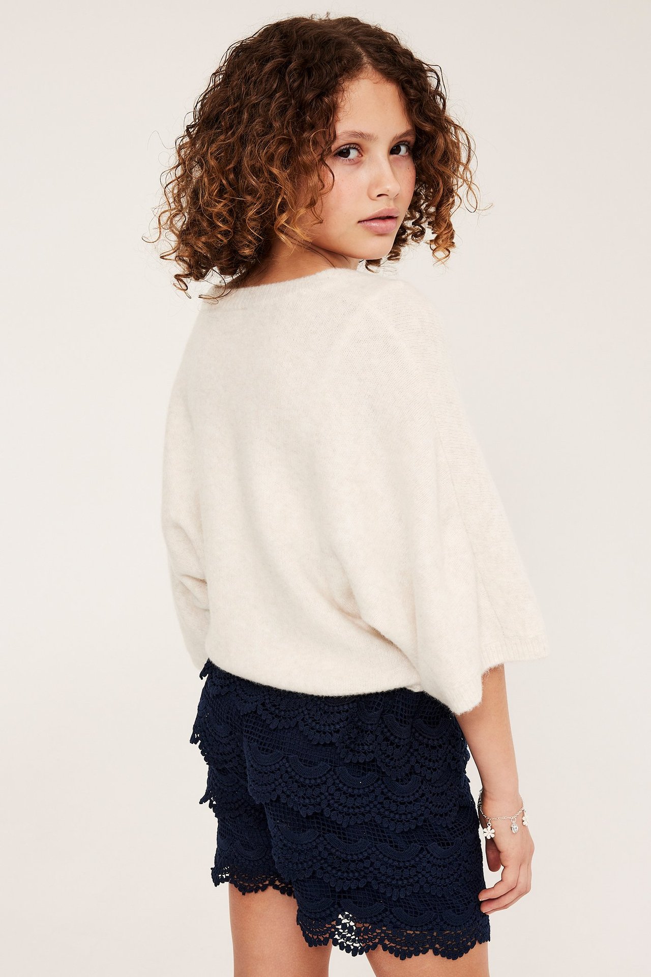 Lapsen takakuva: tummansiniset pitsishortsit, luonnonvalkoinen neulepusero ja rannekoru.