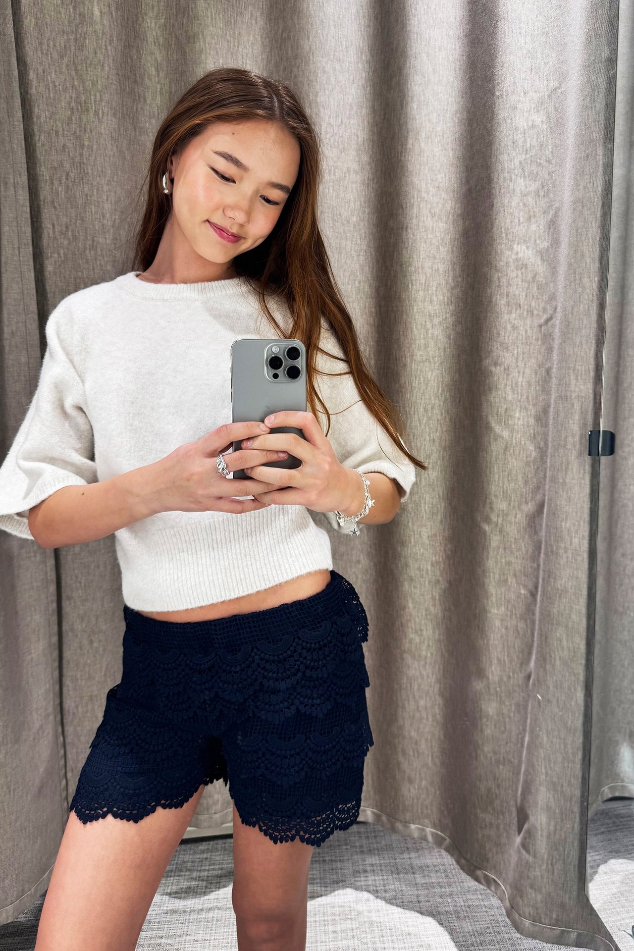 Edestä: Nainen yllään tummansiniset kerrokselliset pitsishortsit ja valkoinen neule, pitäen puhelinta.