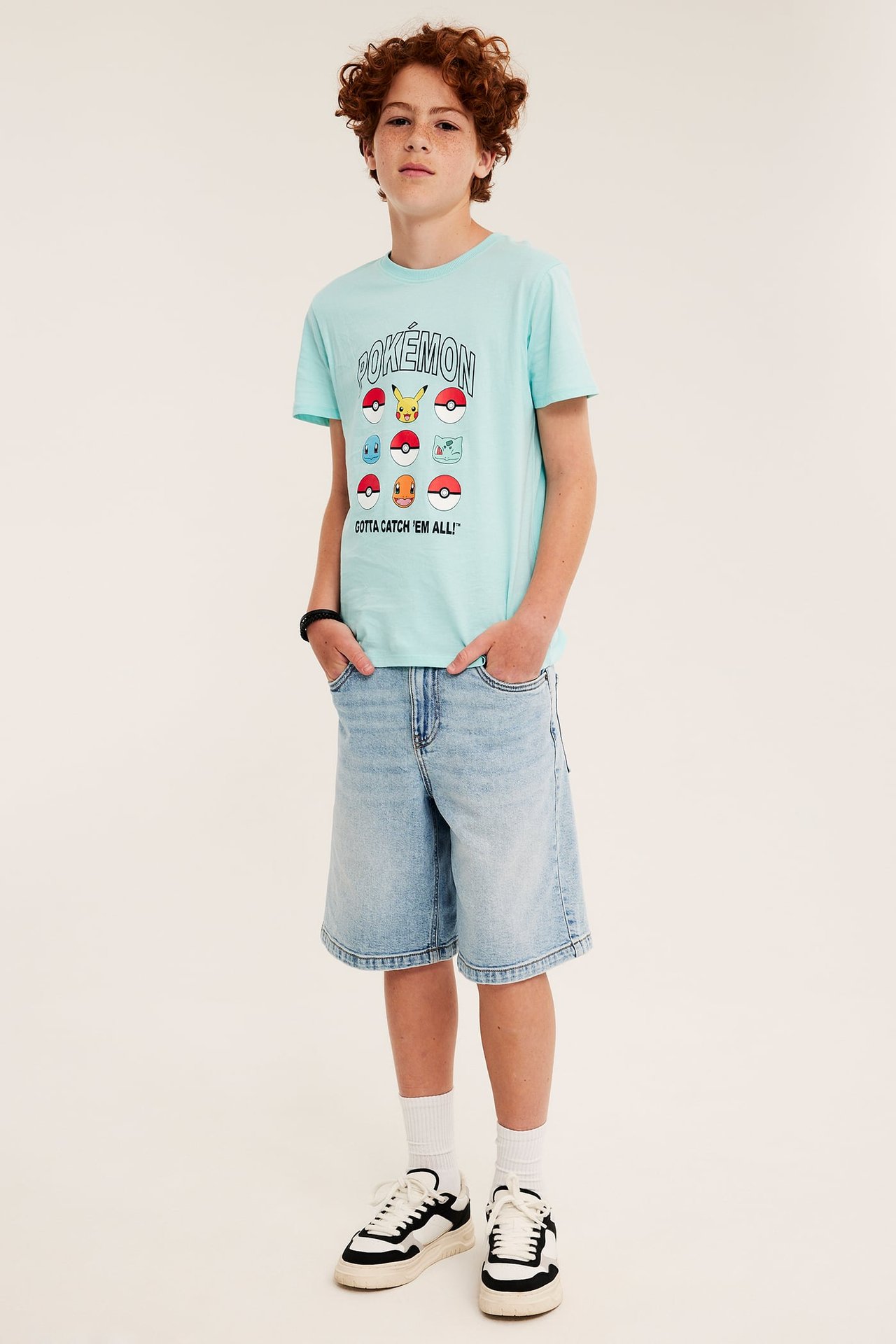 Forfra: Et barn i en lysturkis Pokémon T-skjorte i bomull, lyseblå denimshorts, hvite sokker og sneakers.