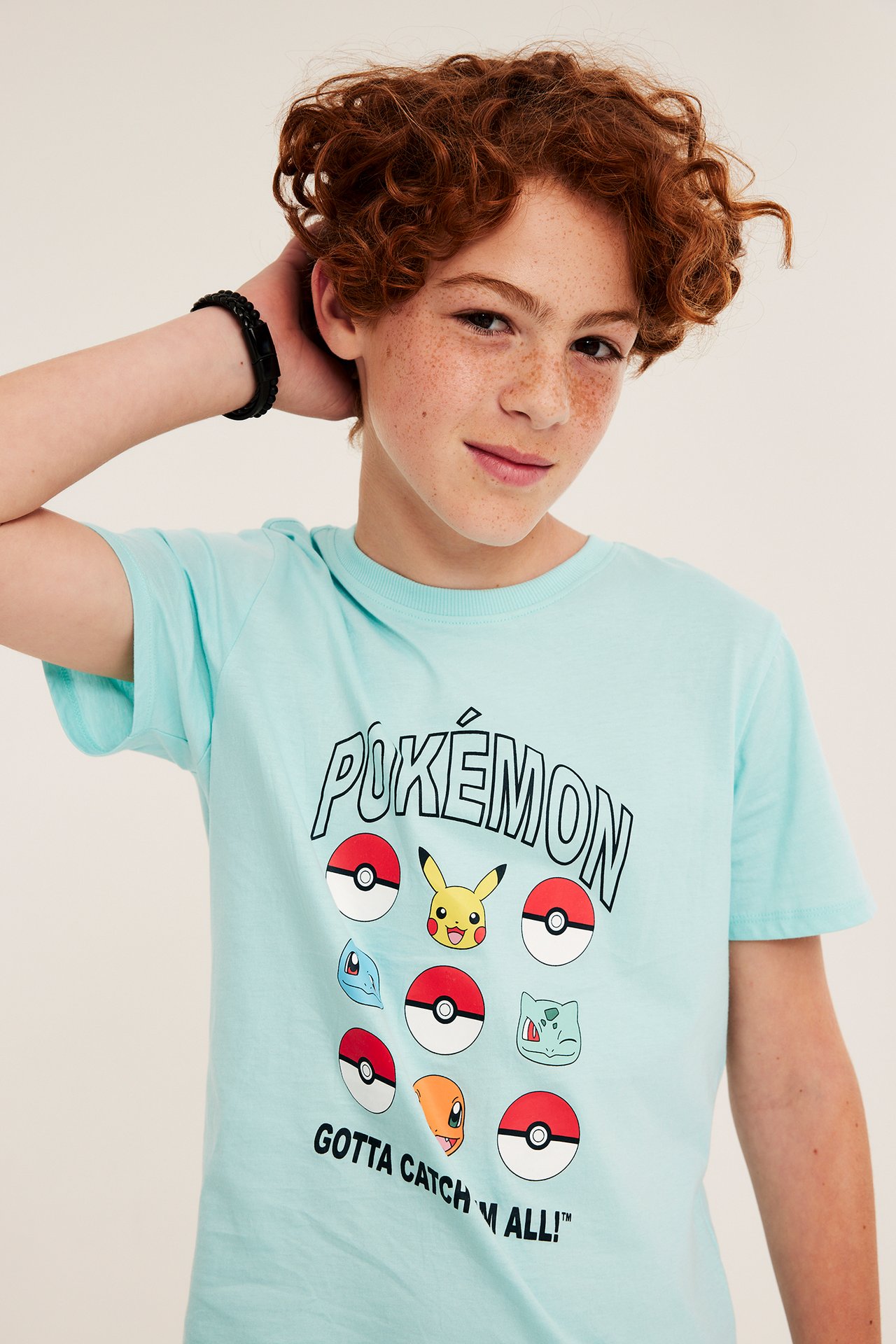 Widok z przodu: dziecko w jasnoturkusowej bawełnianej koszulce z nadrukiem Pokémon i czarna pleciona bransoletka.