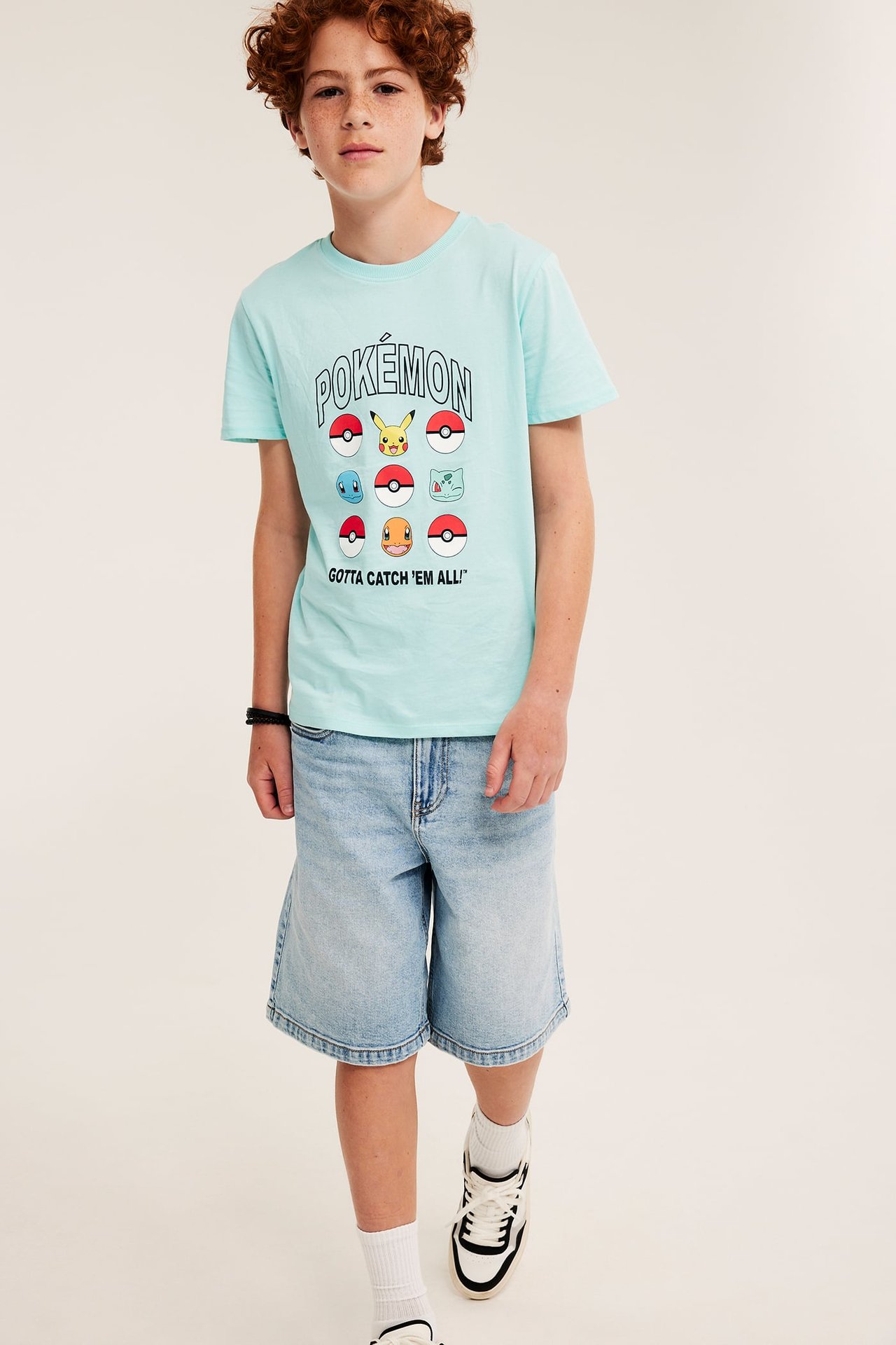 Framifrån: kid i ljust turkos Pokémon-T-shirt i bomull, ljusa jeansshorts och sneakers.