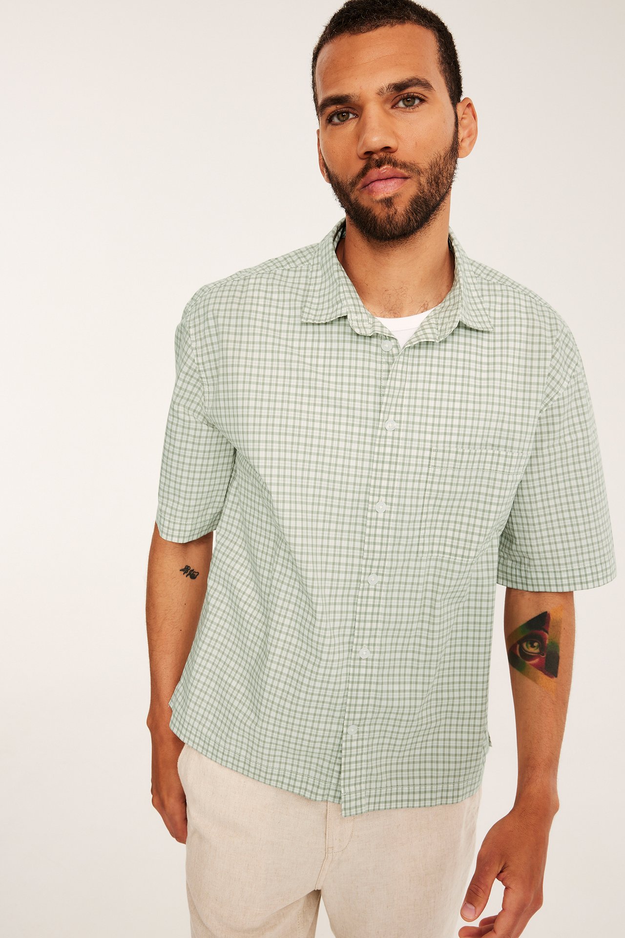 Frontvy av en man i en ljusgrön rutig kortärmad poplinskjorta över en vit t-shirt och ljusbeige byxor.