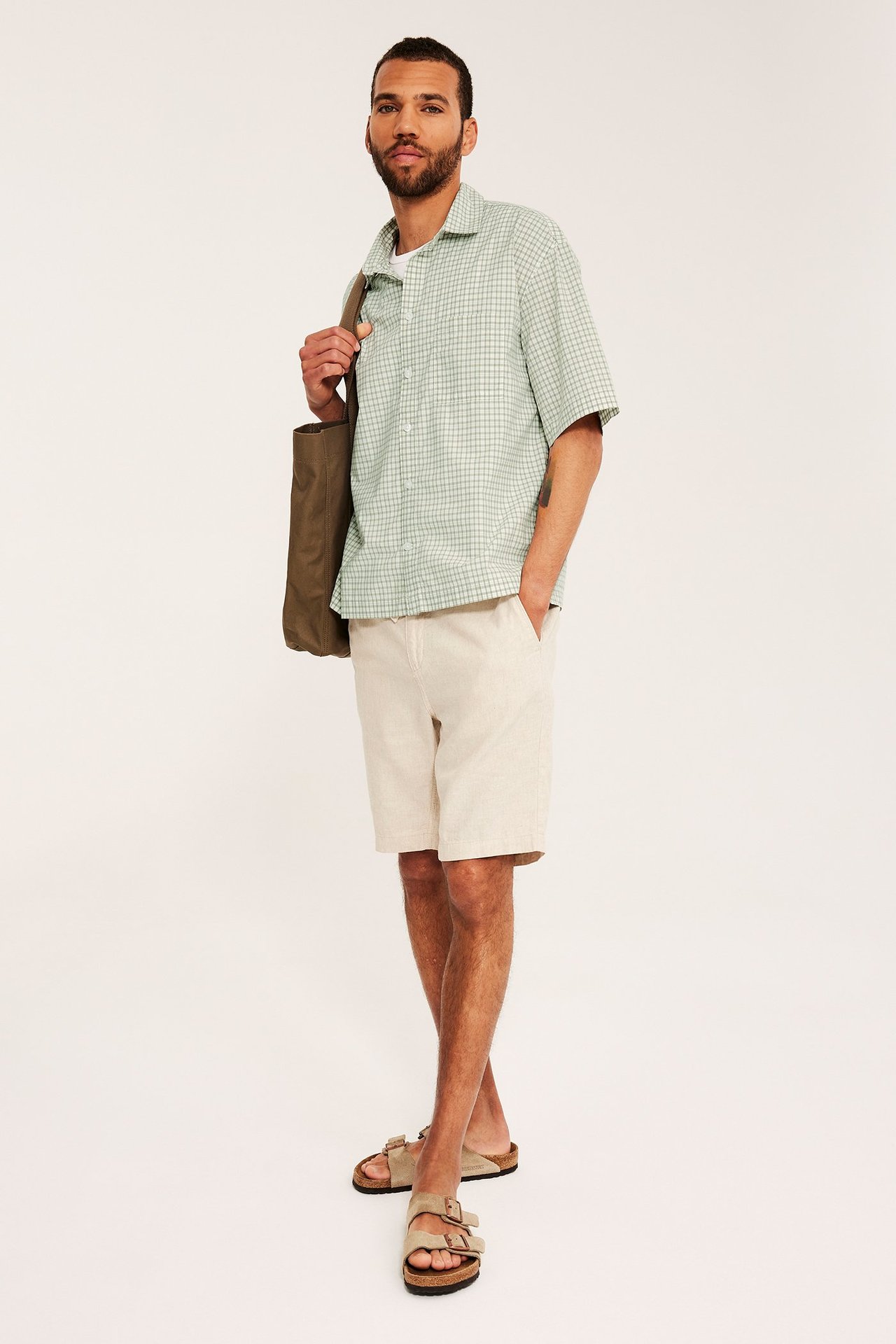 Framifrån: Man i ljusgrön rutig kortärmad poplinskjorta, ljusbeige shorts, beiga sandaler, håller olivgrön tygkasse.