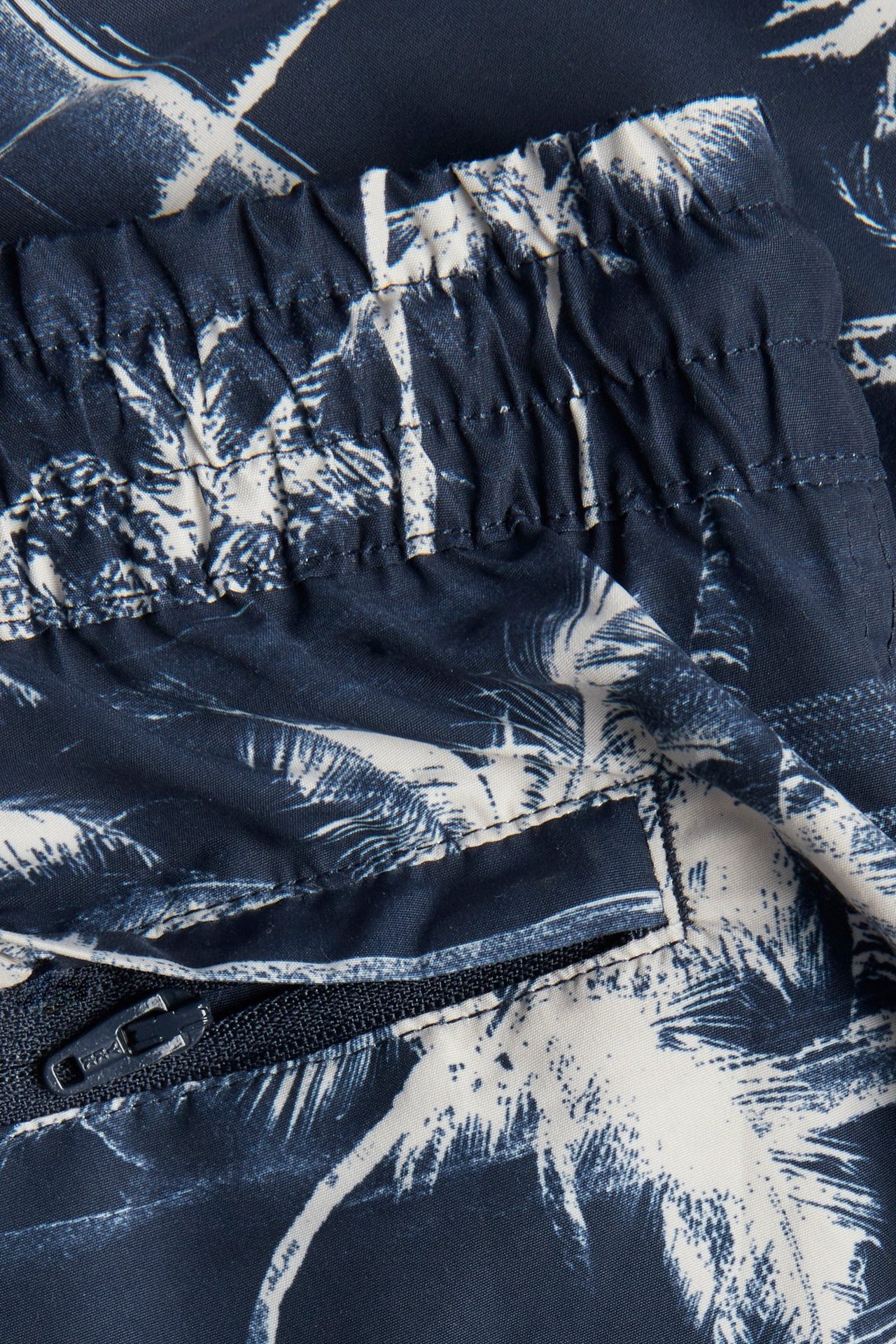 Nærbilde av mørkeblå mønstrede badeshorts med elastisk linning og glidelåslomme.