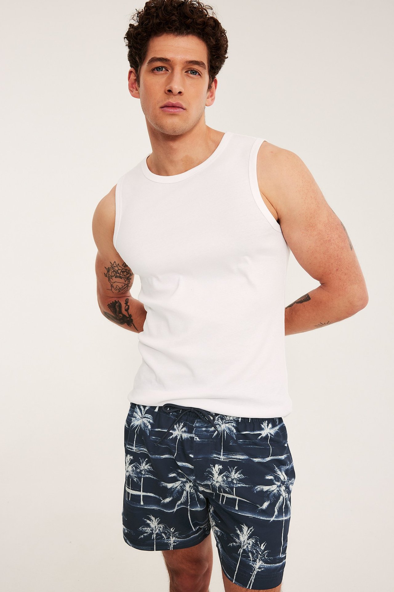 Forfra, mann i mørkeblå mønstrede badeshorts med snøring og en hvit singlet.