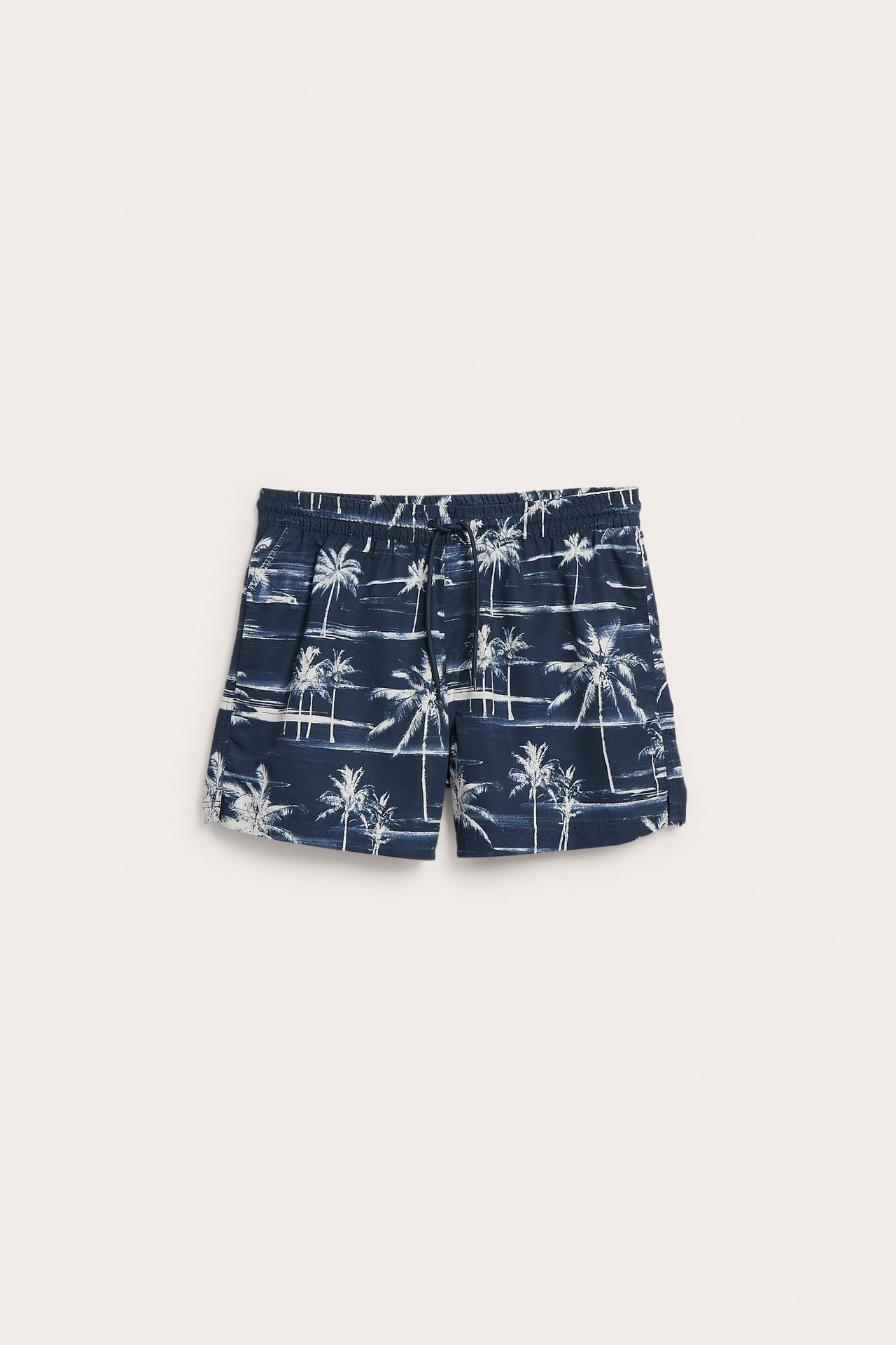 Miesten edestä näkyvät tummansiniset uimashortsit valkoisella palmupuukuvioinnilla, joustava vyötärö ja kiristysnyöri.