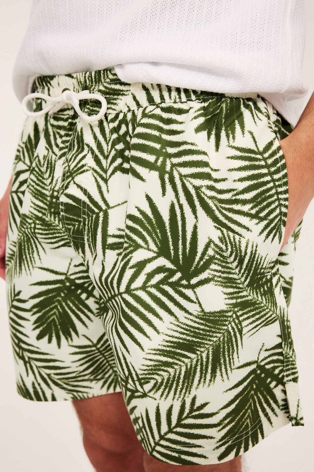Närbild av man i gröna palmönstrade badshorts med vit dragsko. Bär en vit t-shirt.