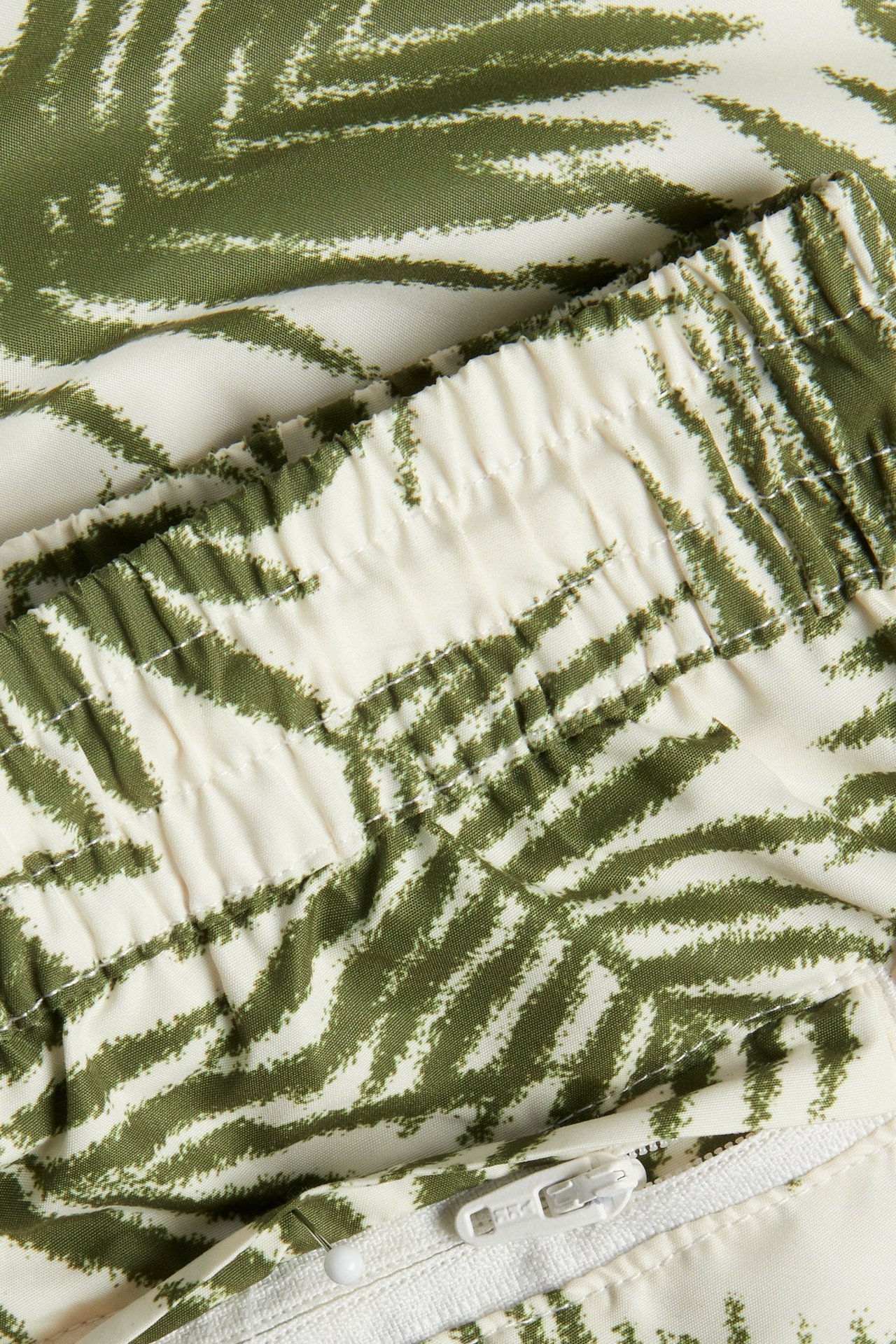 Nærbilde av mønstrede badeshorts for menn med et khakigrønt abstrakt mønster, elastisk linning og glidelås.