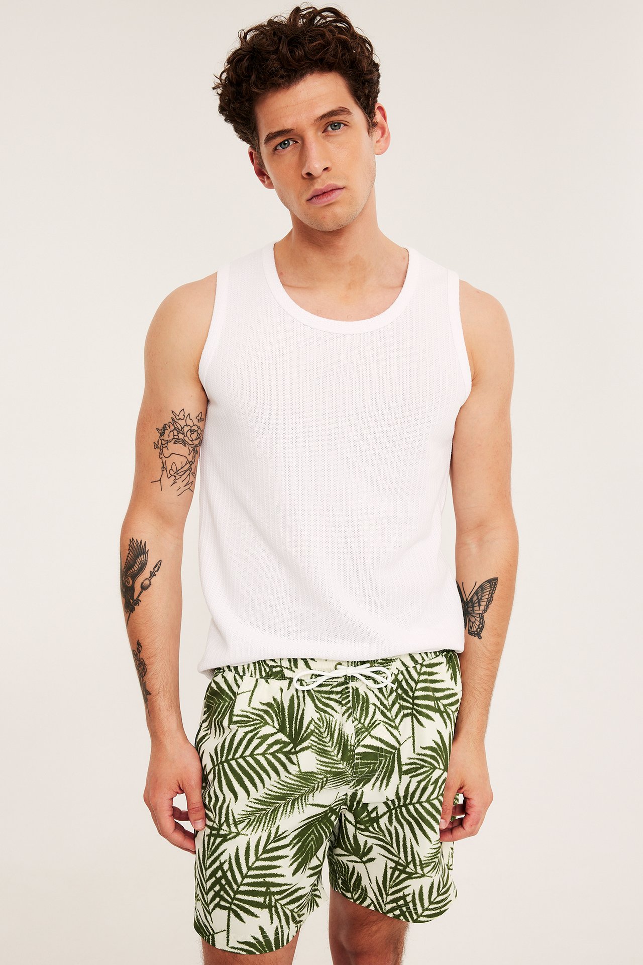 Framifrån: Man i gröna palmbladsmönstrade badshorts med dragsko i midjan och ett vitt linne.