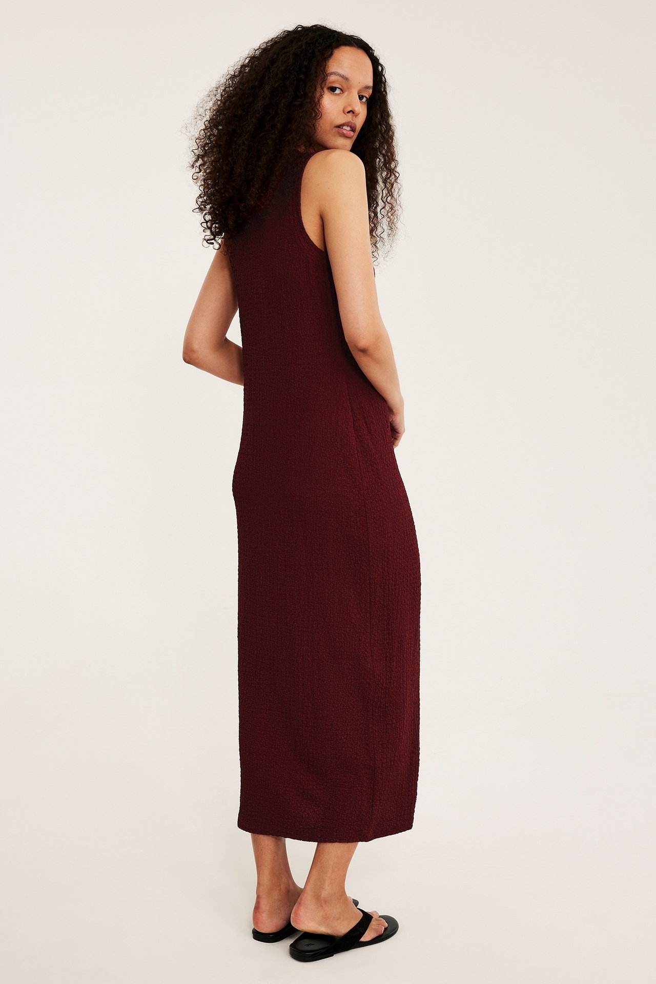 Naisen sivukuva yllään punainen strukturoitu hihaton midimekko ja mustat litteät sandaalit.