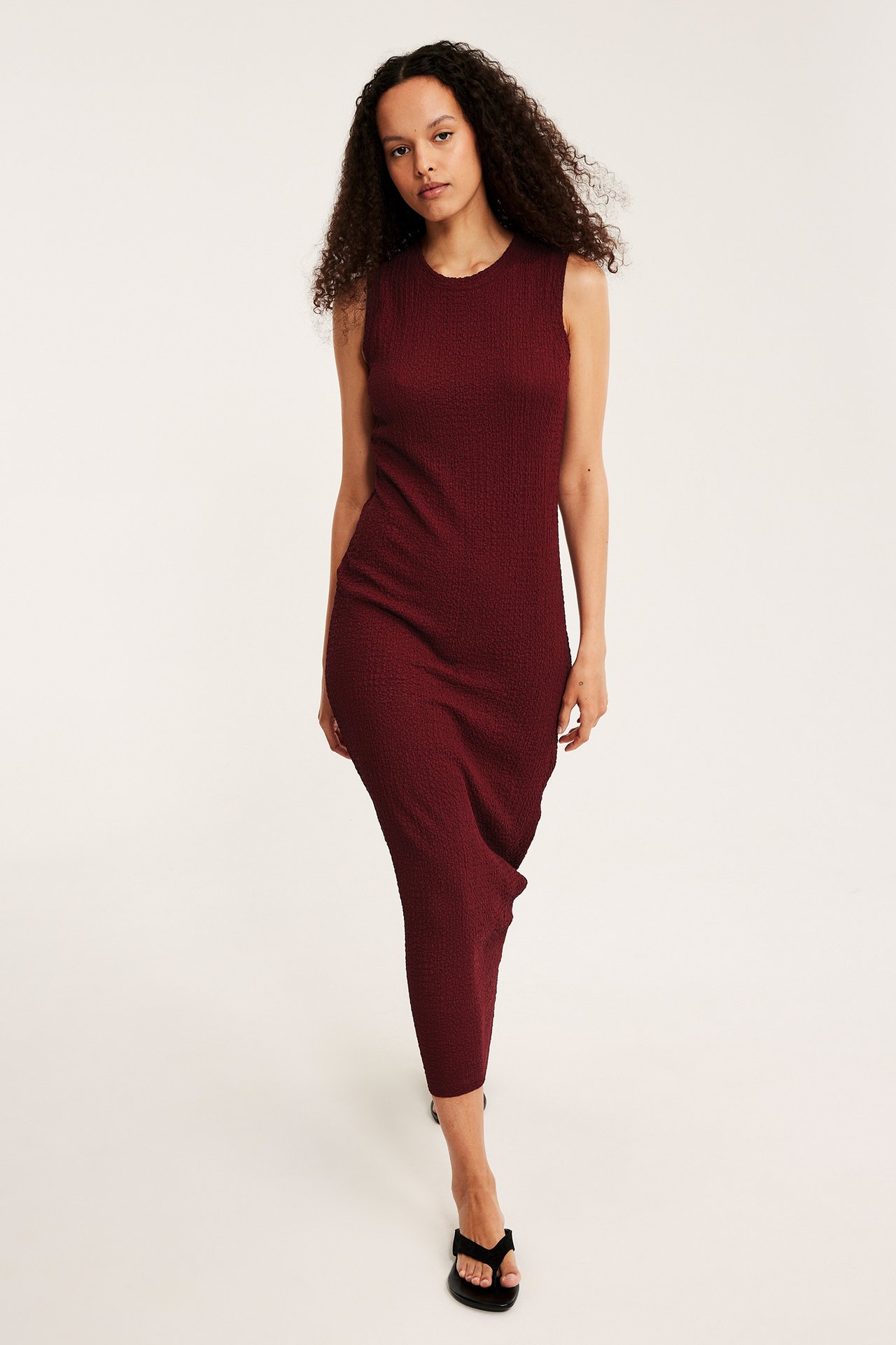 Frontvy av kvinna i figurnära mörkröd strukturerad ärmlös midiklänning och svarta sandaler.