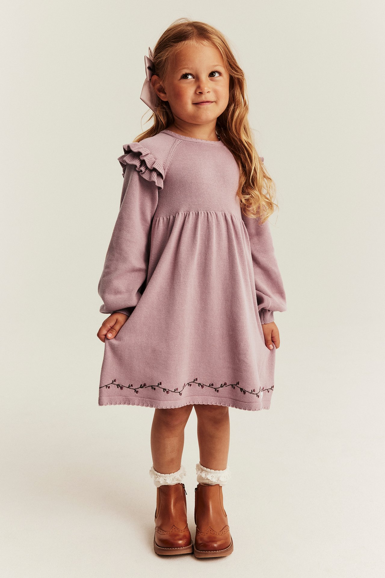 Frontvy: Barn i mauve volangstickad klänning, broderad fåll, bruna stövlar, vita strumpor, rosa rosett.