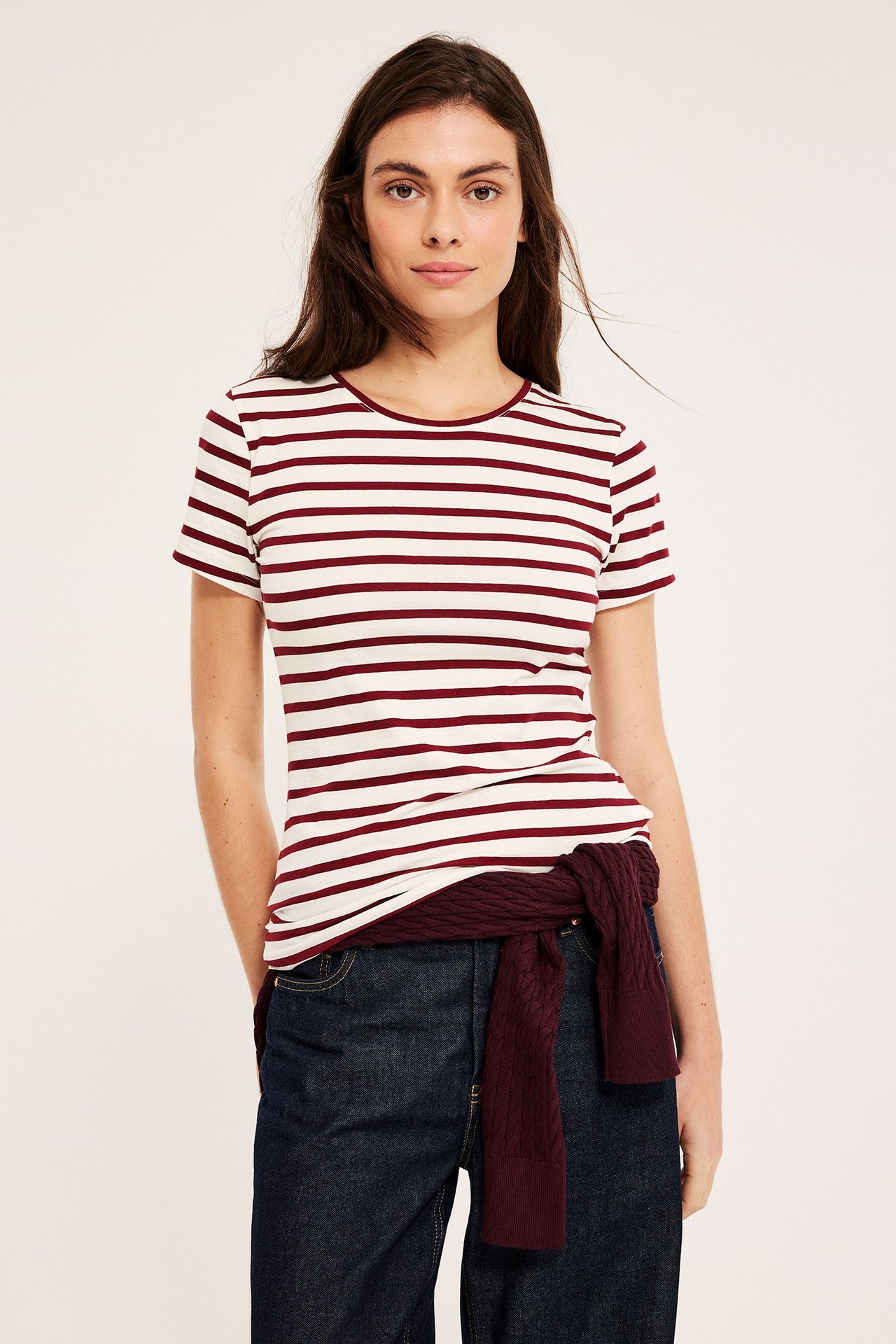 Widok z przodu: kobieta w czerwono-białym pasiastym topie, ciemnych jeansach, bordowym swetrze w pasie.