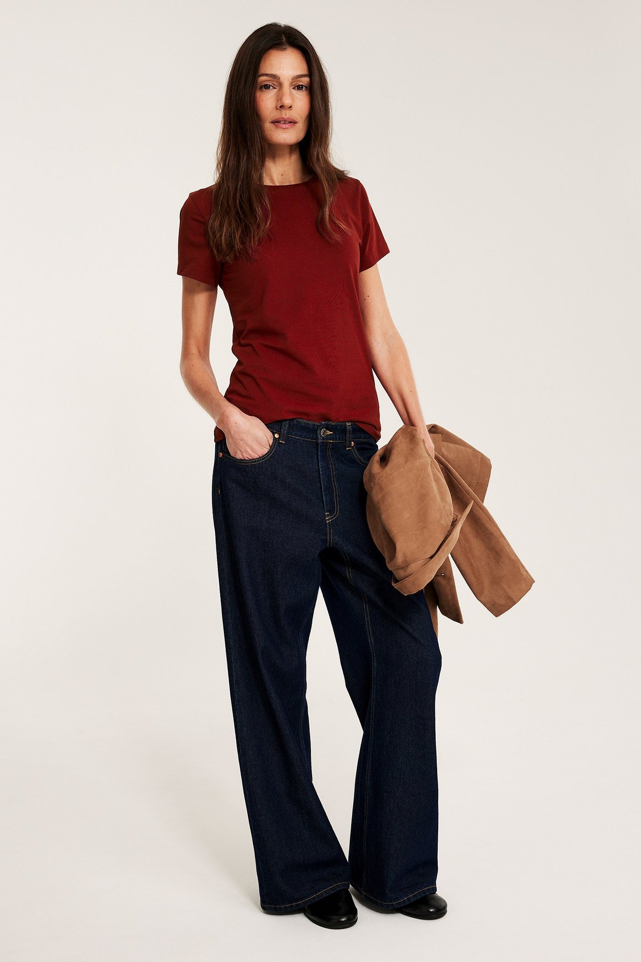 Frontbilde: kvinne i rød kortermet basetopp, mørkeblå vide jeans, holder en brun jakke.