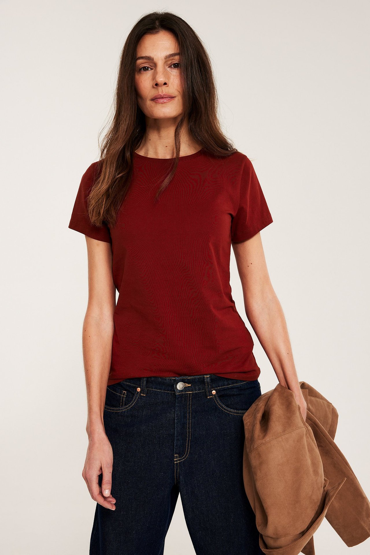 Framfra, kvinne i ensfarget mørkerød kortermet basetopp, mørkeblå jeans og holder en brun jakke.
