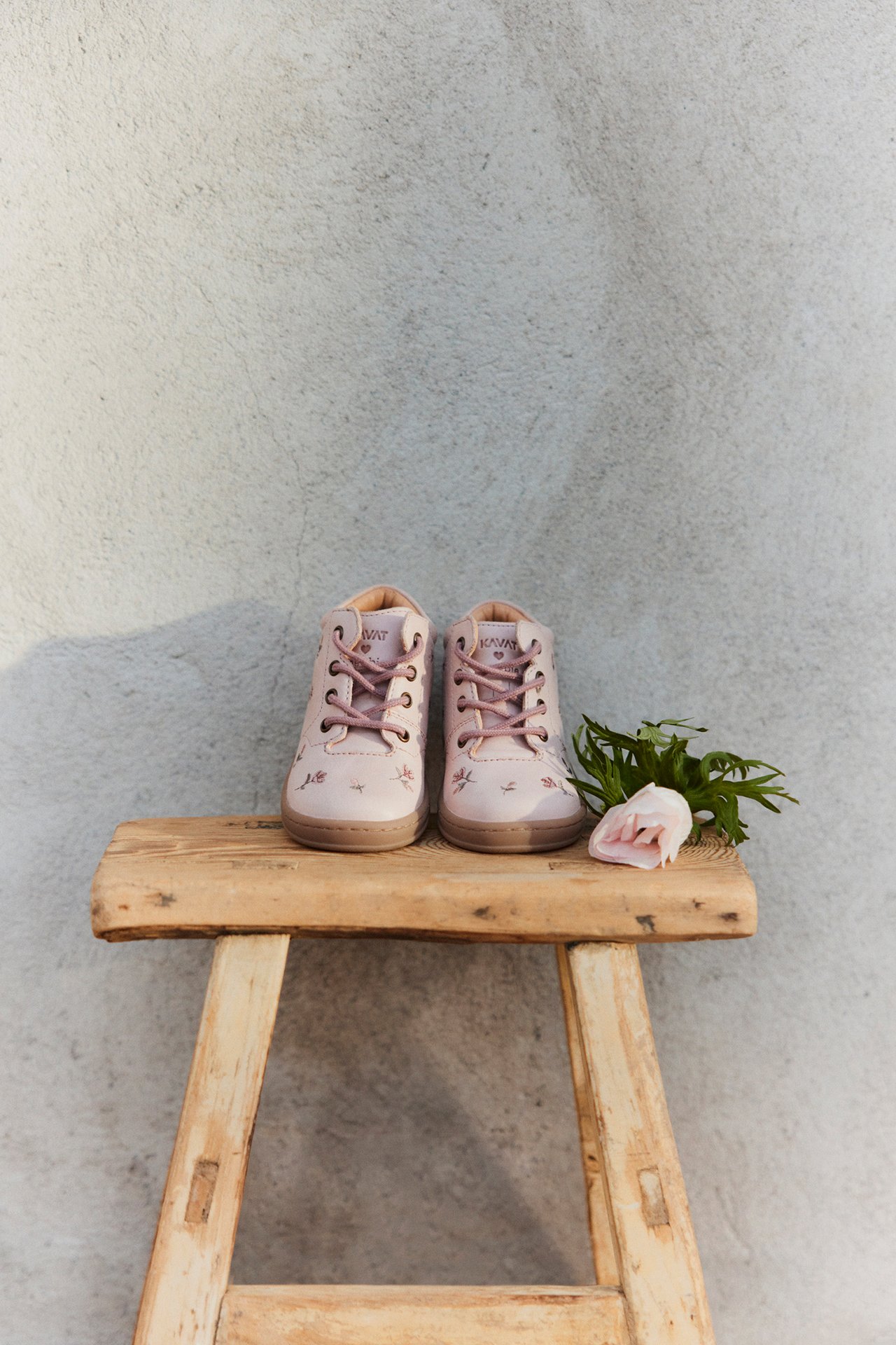 Edestäpäin vaaleanpunaiset lasten kukkakuosiset nauhakengät puujakkaralla vaaleanpunaisen kukan vieressä.