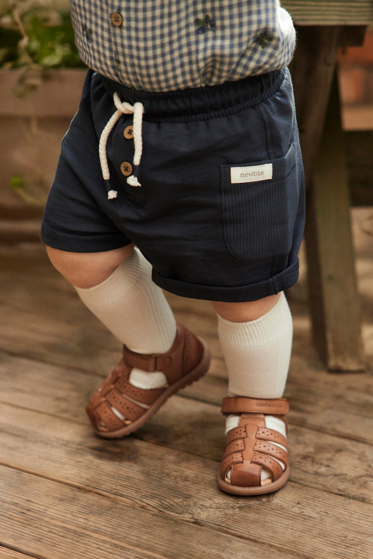 Närbild på baby som bär bruna lädersandaler, mörkblå shorts, en blårutig skjorta och vita strumpor.