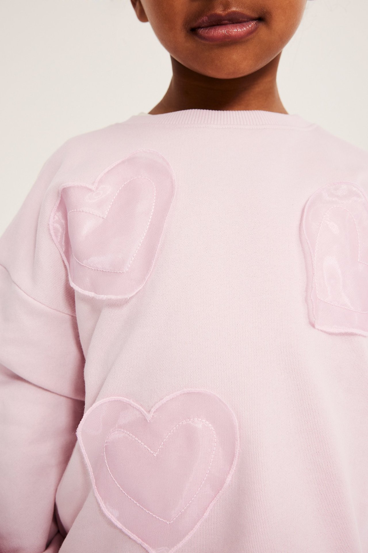 Zbliżenie przodu dziecka w jasnoróżowej bluzie z mieszanki bawełny z aplikacjami z serc z organzy i ściągaczem.