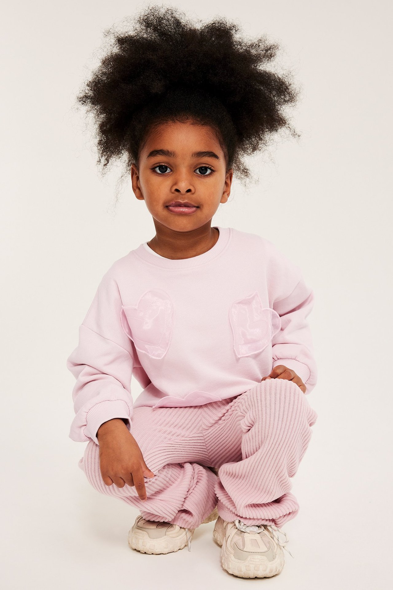 Edestä kuvattu kyykistyvä lapsi vaaleanpunaisessa collegepuserossa organzahjertasovelluksin, housuissa ja tennareissa.