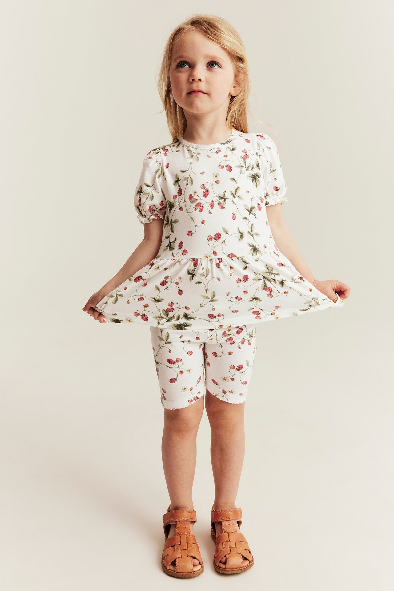 Framsida av barn i offwhite peplumtopp med smultronmönster, puffärm, matchande shorts och bruna sandaler.