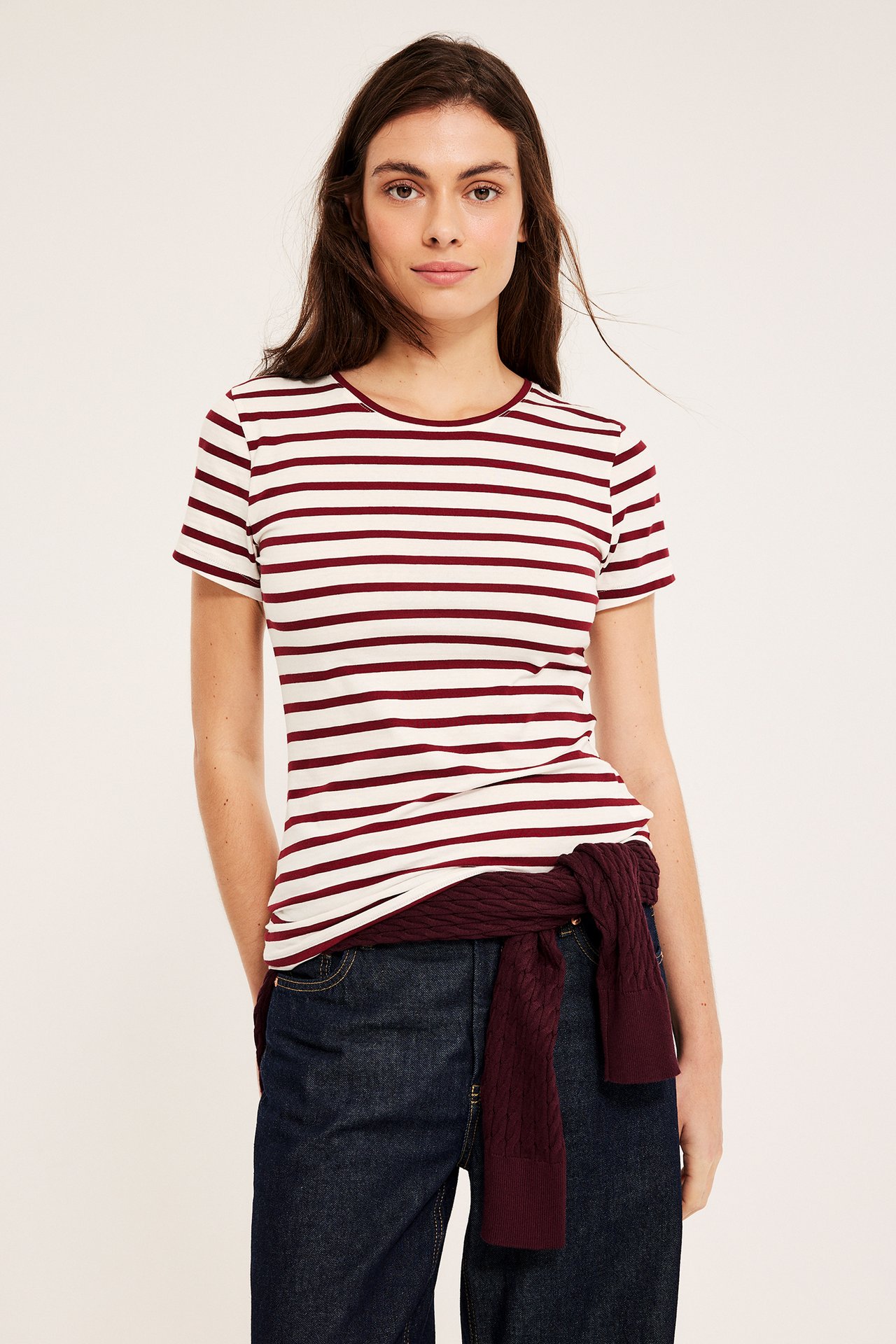 Frontutsikt: kvinne i hvit-mørkerød stripet kortermet topp, mørkeblå jeans, burgunder genser knyttet rundt midjen.
